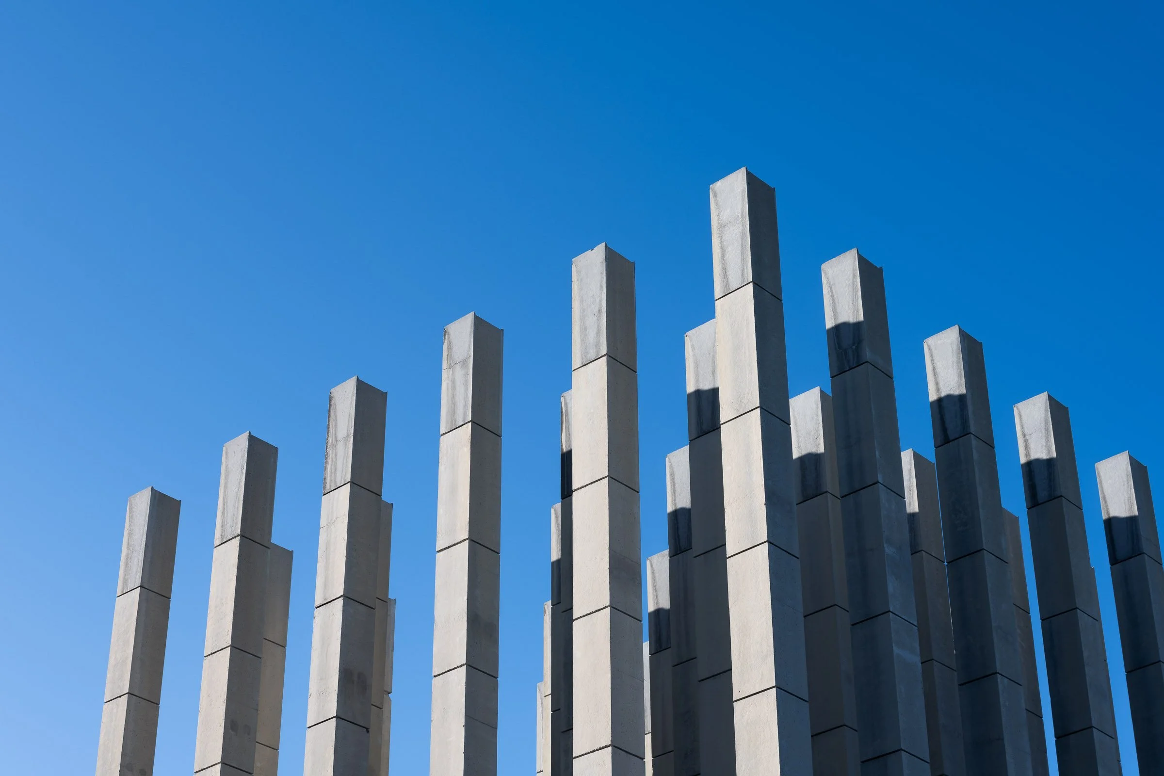A row of tall, rectangular concrete pillars with shadows cast on them under a clear blue sky.