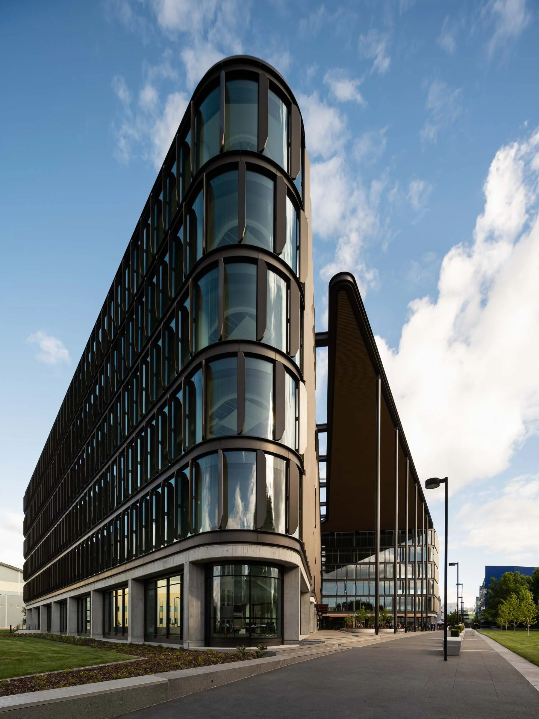 Modern multi-story building with rounded glass windows and black framing, set against a partly cloudy sky and a landscaped sidewalk.