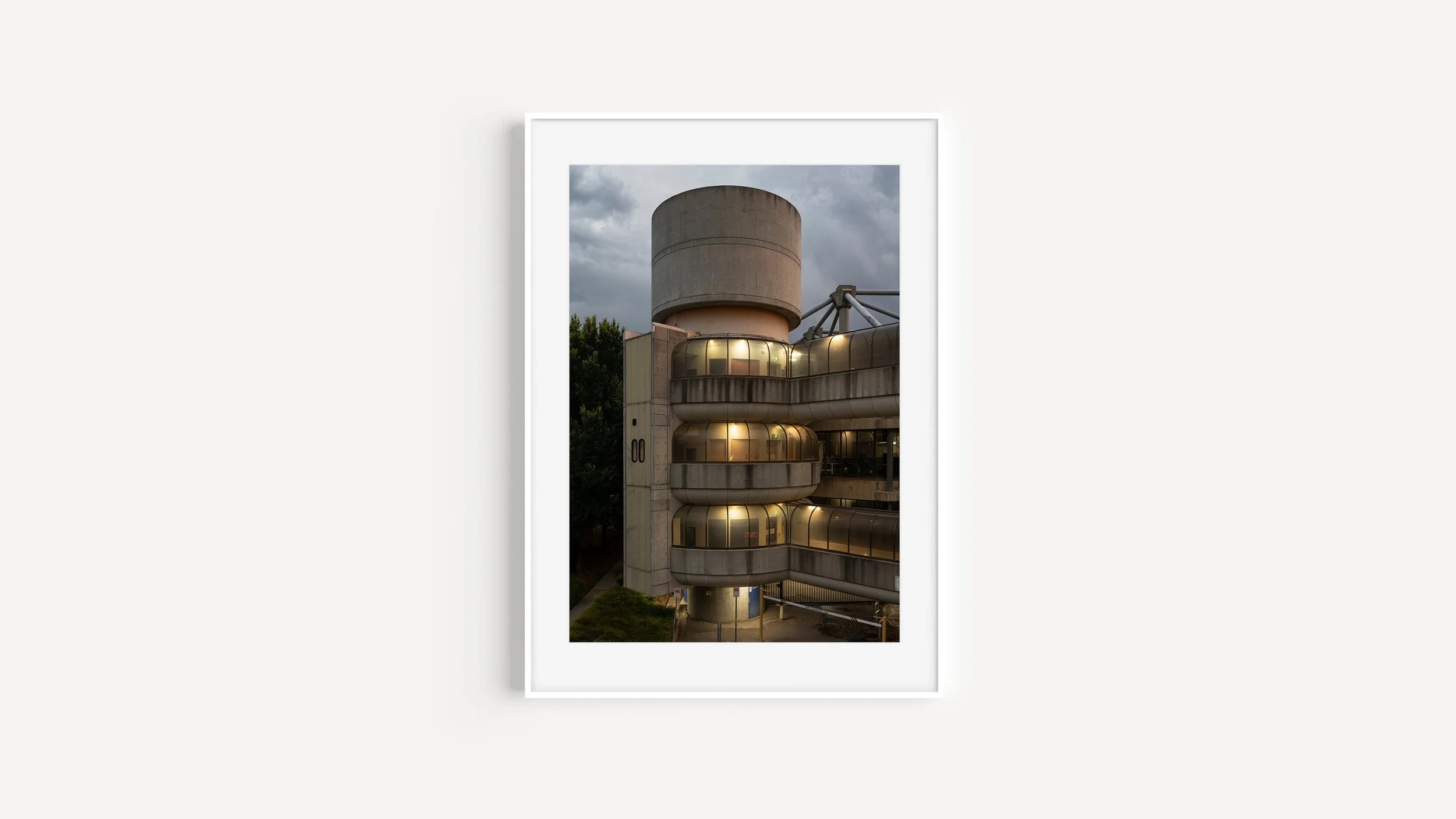 The image depicts a modern building with rounded balconies and large glass windows, a tower-like structure with a concrete exterior, situated outdoors with trees and a cloudy sky background.