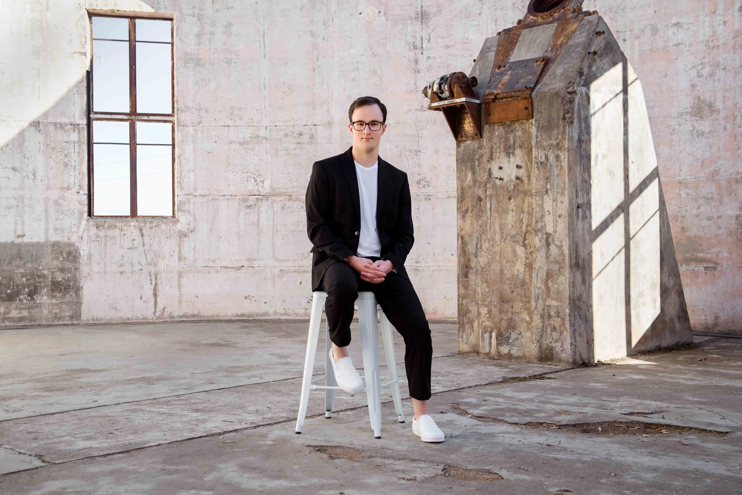 A young man with dark hair, glasses, wearing a black blazer, white t-shirt, black pants, and white sneakers, sitting on a white stool in an industrial space with a textured concrete wall and window, with a rusty industrial machine on a pedestal nearby.