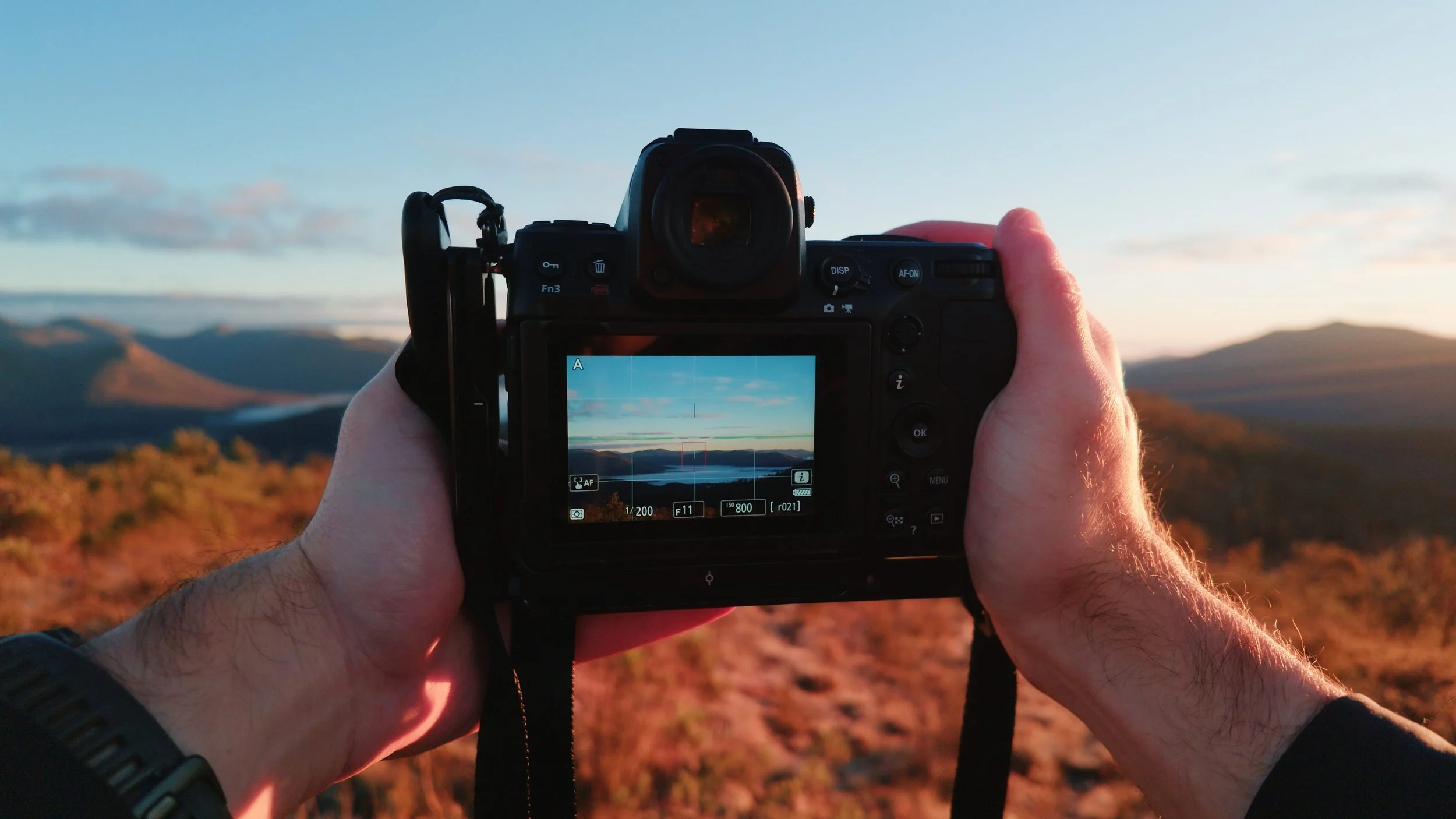 Person taking a photograph of a scenic landscape with a digital camera during sunset or sunrise in a mountainous area.
