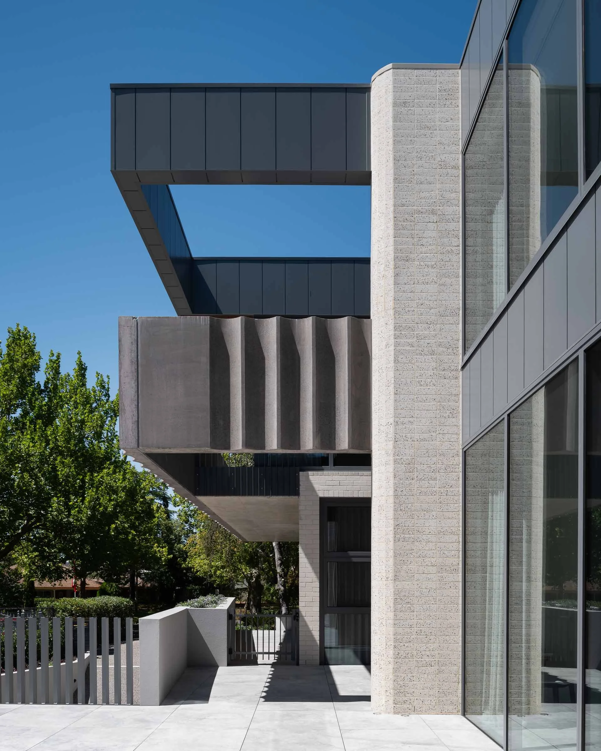 Modern building exterior with glass windows, textured white brick wall, gray steel panels, concrete fence, and trees in background under clear blue sky.