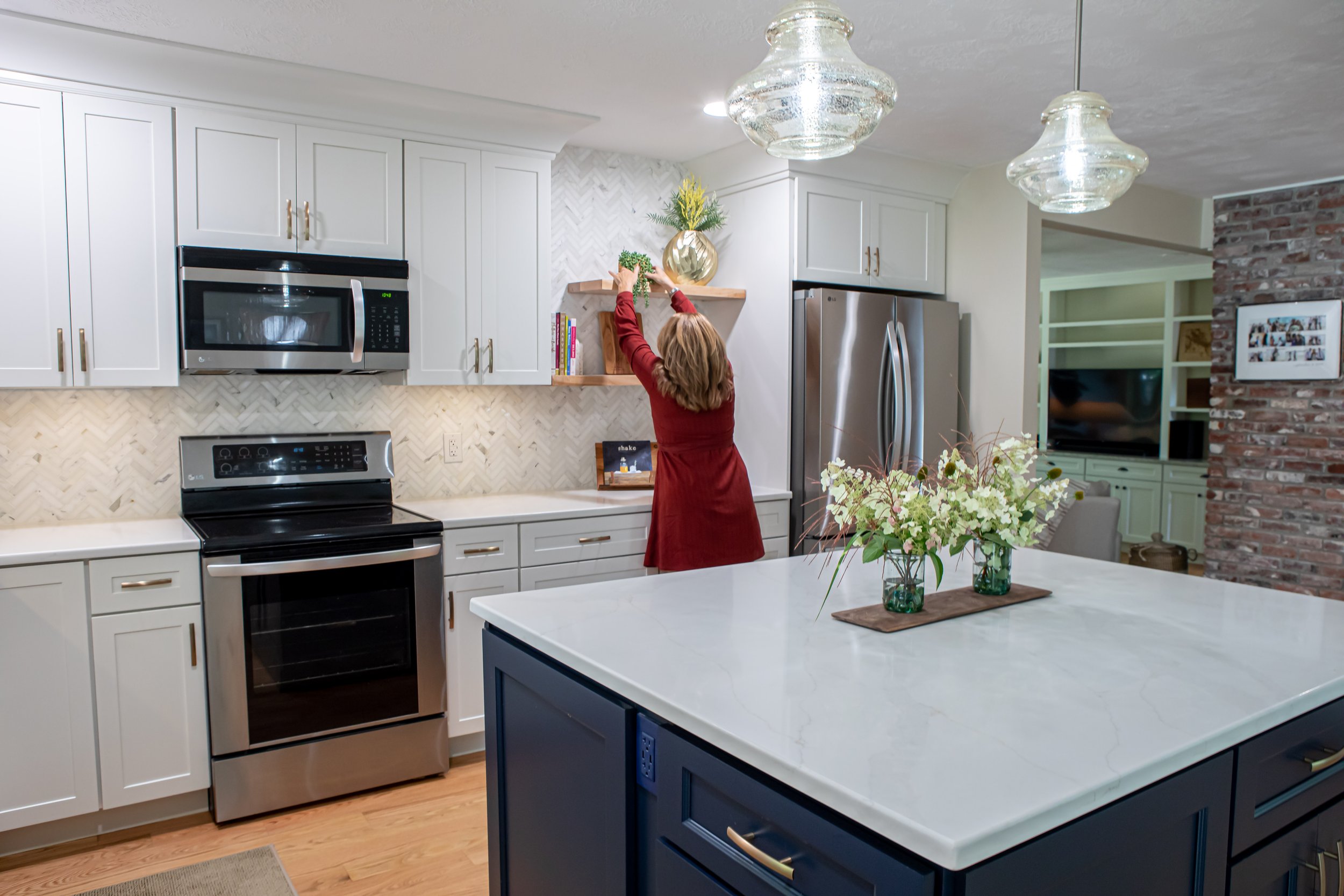 Anne Guyotte placing a decorative item on a wooden shelf in a modern kitchen with white cabinets, stainless steel appliances, and a large island with a vase of flowers.