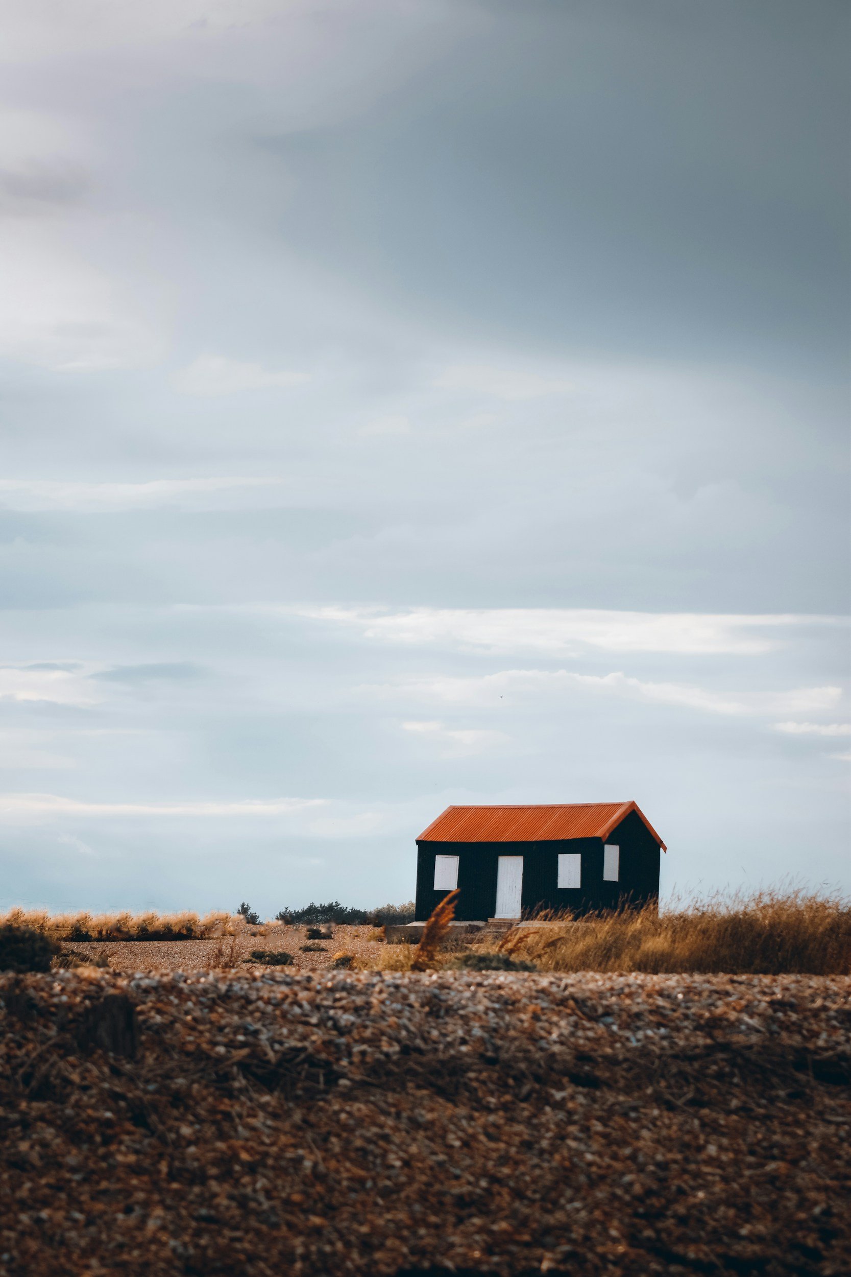 A small house with a red roof and black walls with white windows sits alone in a field of dry grass, under a cloudy sky.