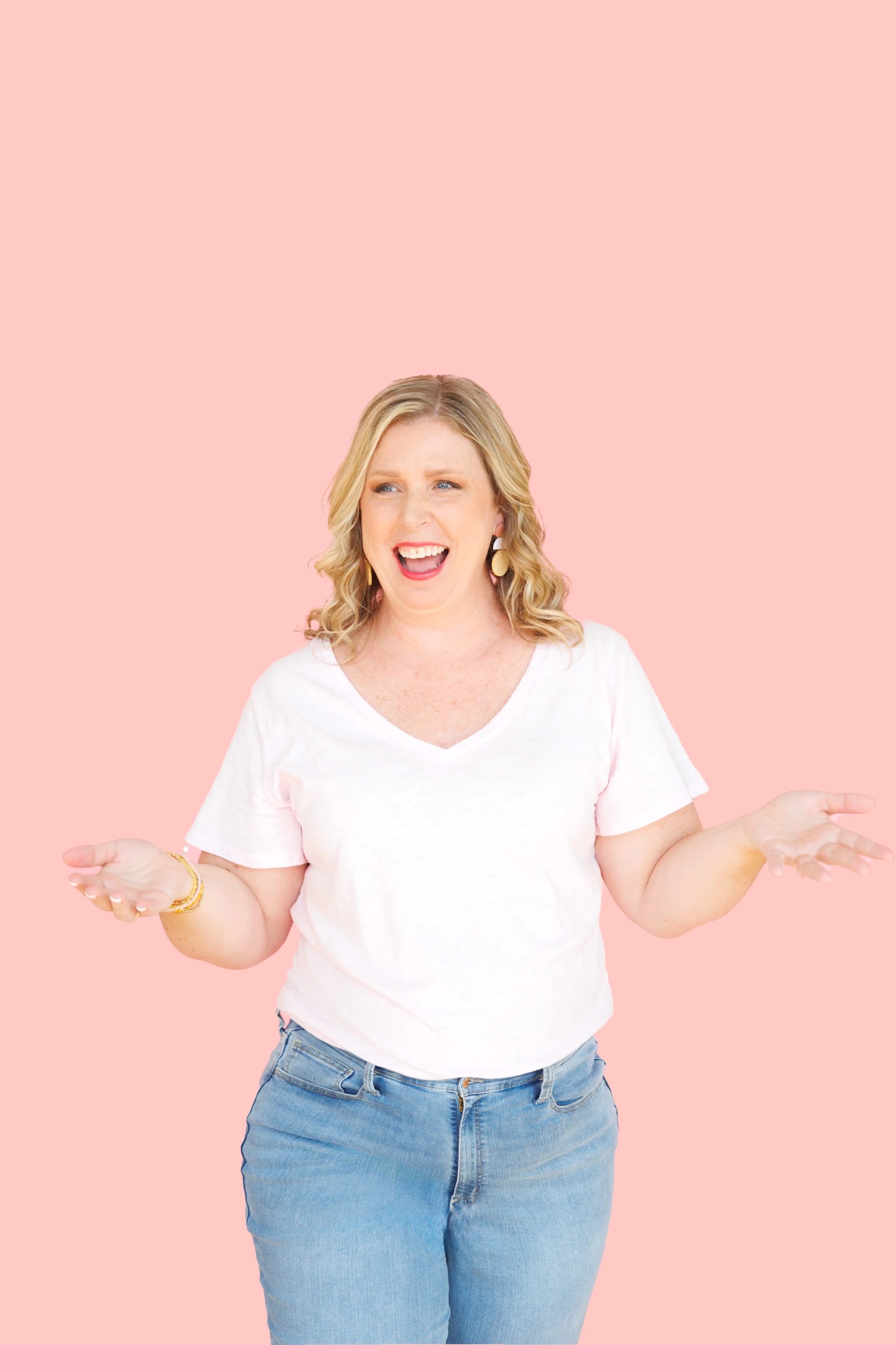 Woman with blonde hair, wearing a white t-shirt and jeans, standing against a pink background with a confused or questioning expression, gesturing with open hands.