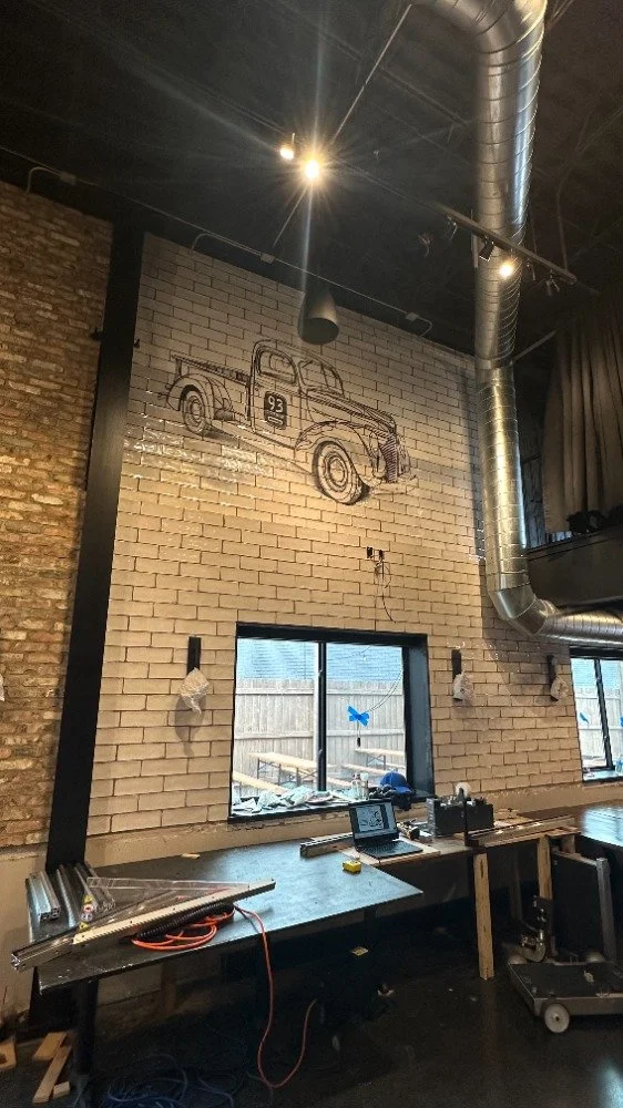Interior of a brewery building under construction with ceramic and brick walls, a large industrial ventilation duct, and two windows. A drawing of a vintage pickup truck with the number 93 on its door is mounted on the wall above the windows.