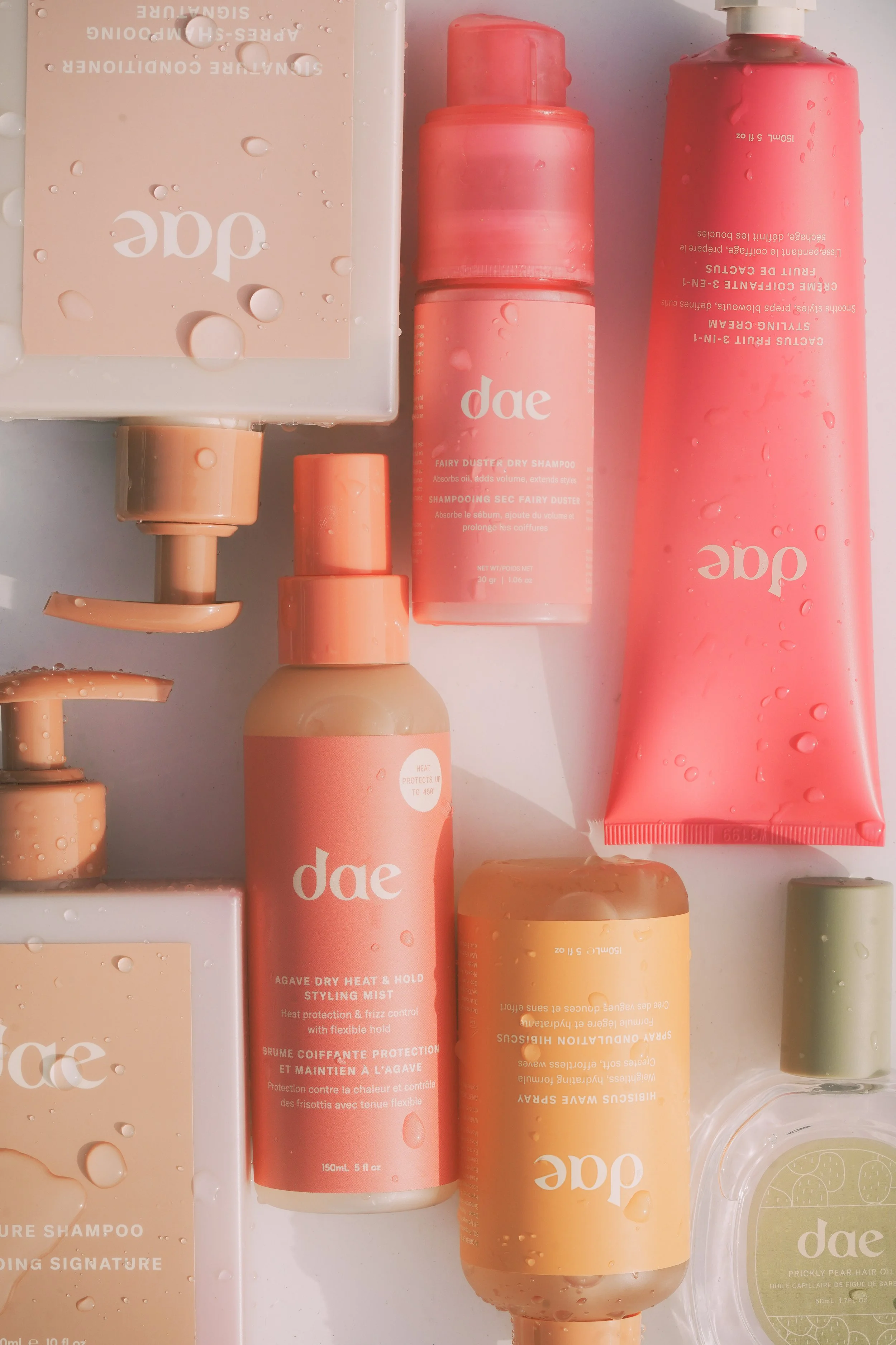 Arrangement of pink and peach Dae hair and styling products, including shampoo, conditioner, mist, and hair oil bottles, with water droplets on packaging.