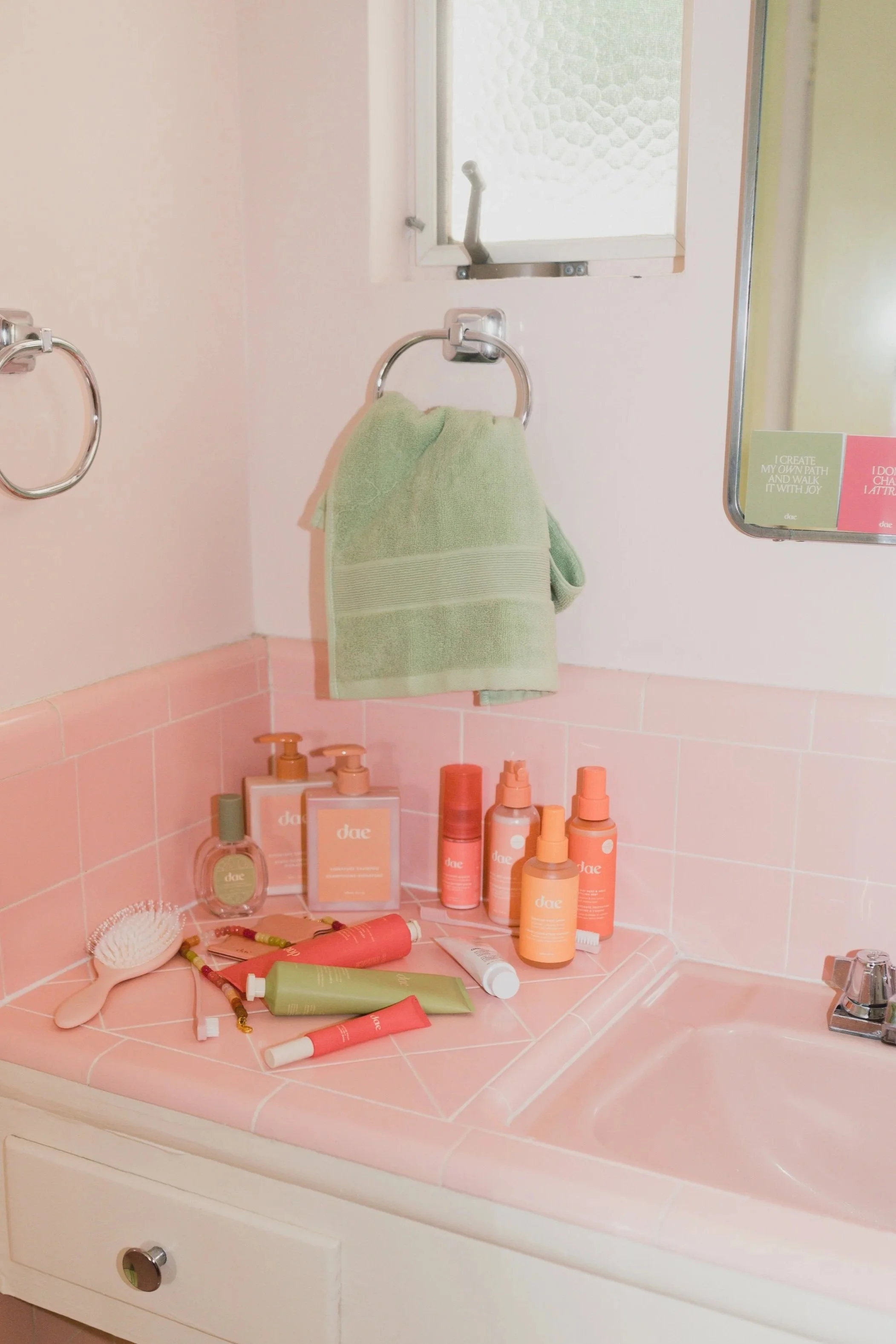 Bathroom sink with pink tiles and various peach-colored skincare products, a pink hairbrush, and a green towel hanging on a towel ring. There is a small mirror and a window above the sink.
