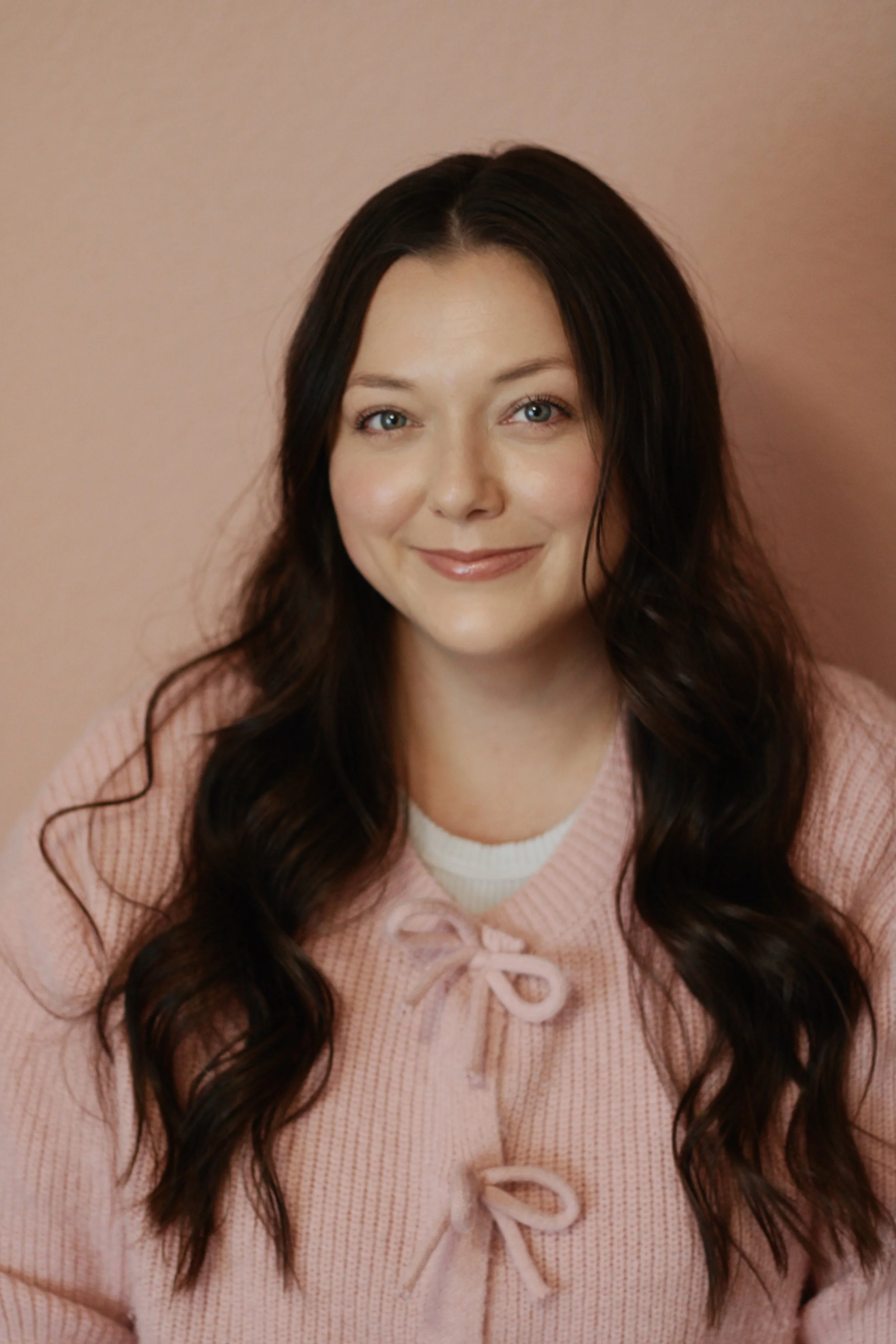 Portrait of a young woman with long, wavy dark hair, wearing a pink sweater with bows, smiling against a pink background.