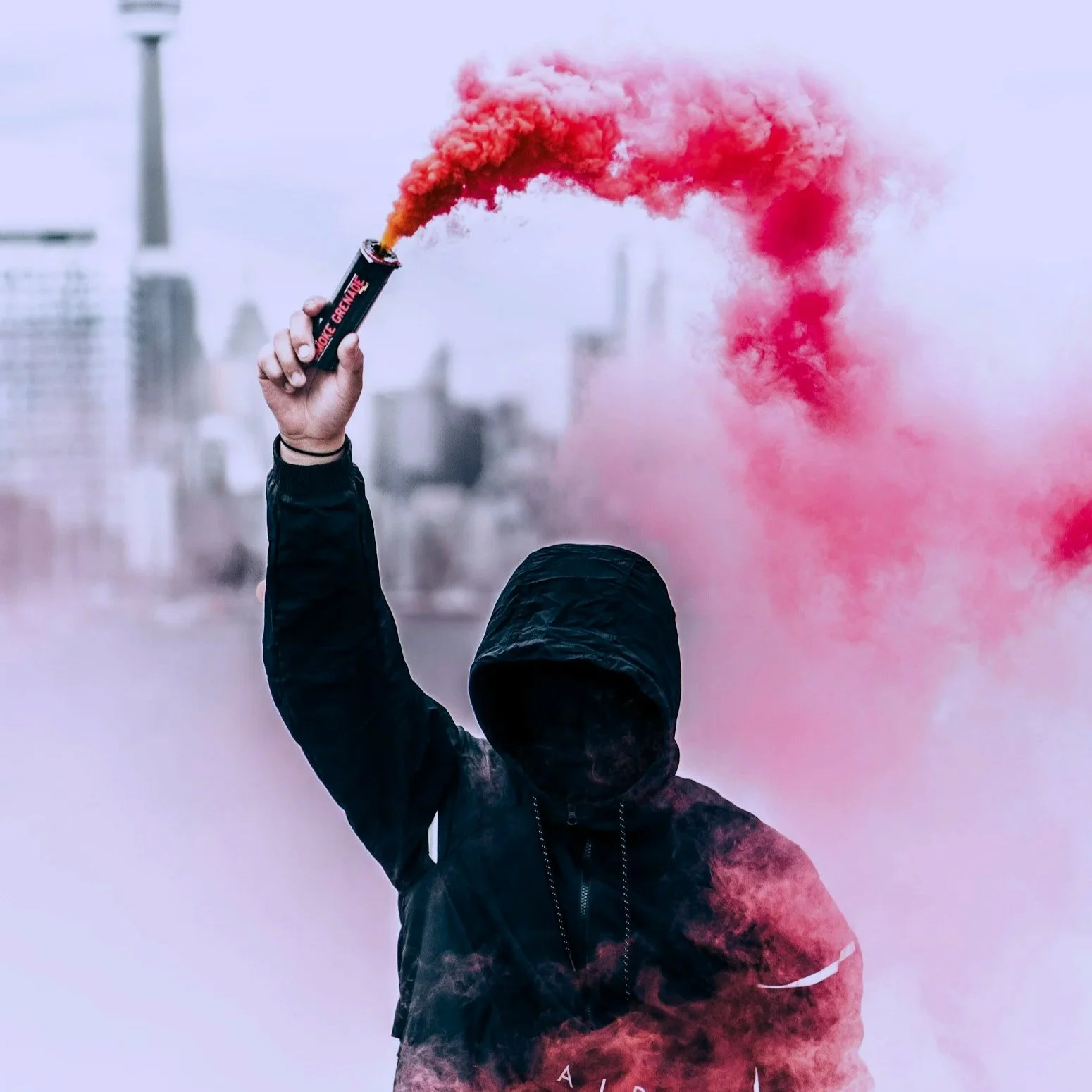 A figure in a hoodie holds up an active smoke grenade as red smoke spews out. The figure's face is not visible beneath the hood.