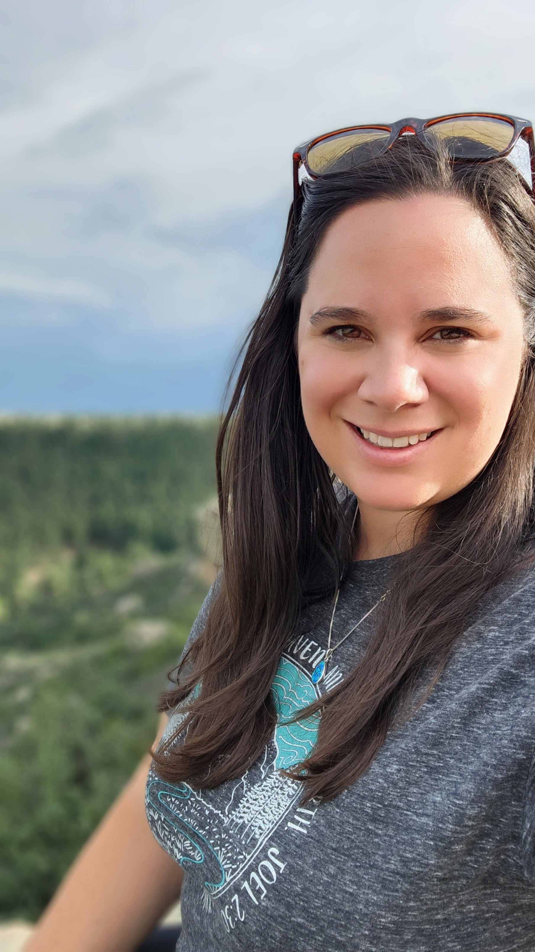 A woman smiling outdoors with hills and a cloudy sky in the background, wearing sunglasses on her head and a gray graphic T-shirt.