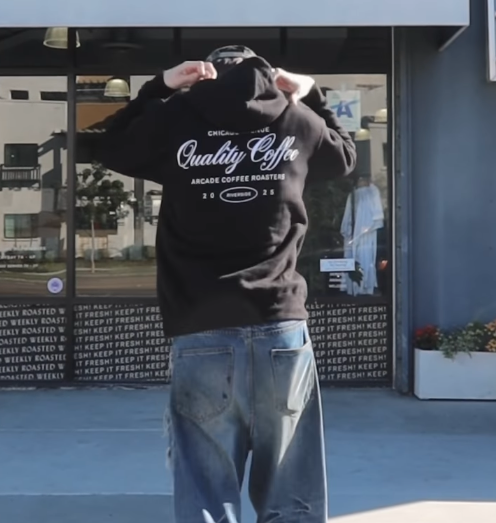 Person standing outside a coffee shop, wearing a black hoodie with white text and jeans, facing away from the camera.