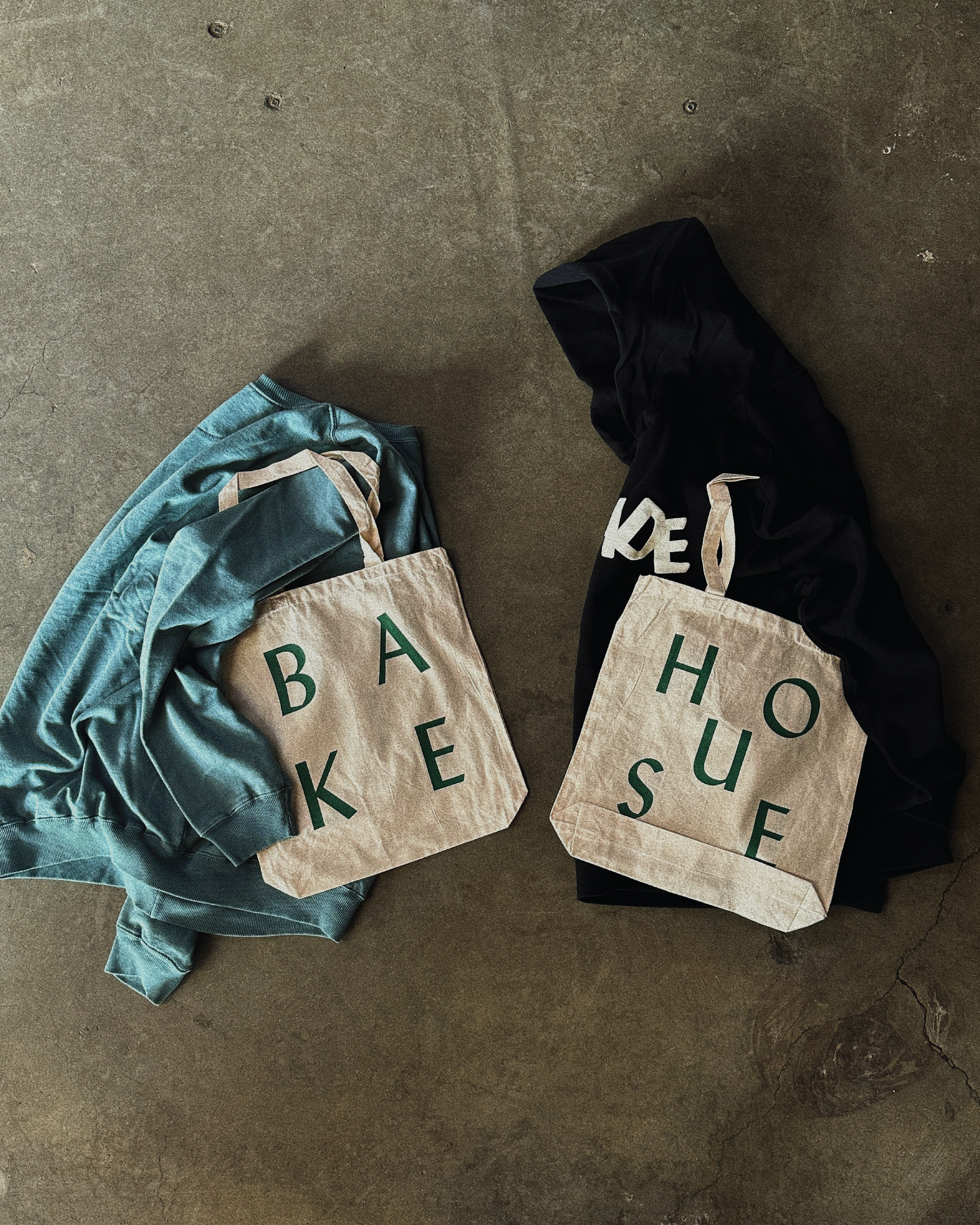 Two cloth tote bags on a concrete floor, one with the words 'BAKE' on it and the other with the words 'HOUSE' on it, each surrounded by clothes.