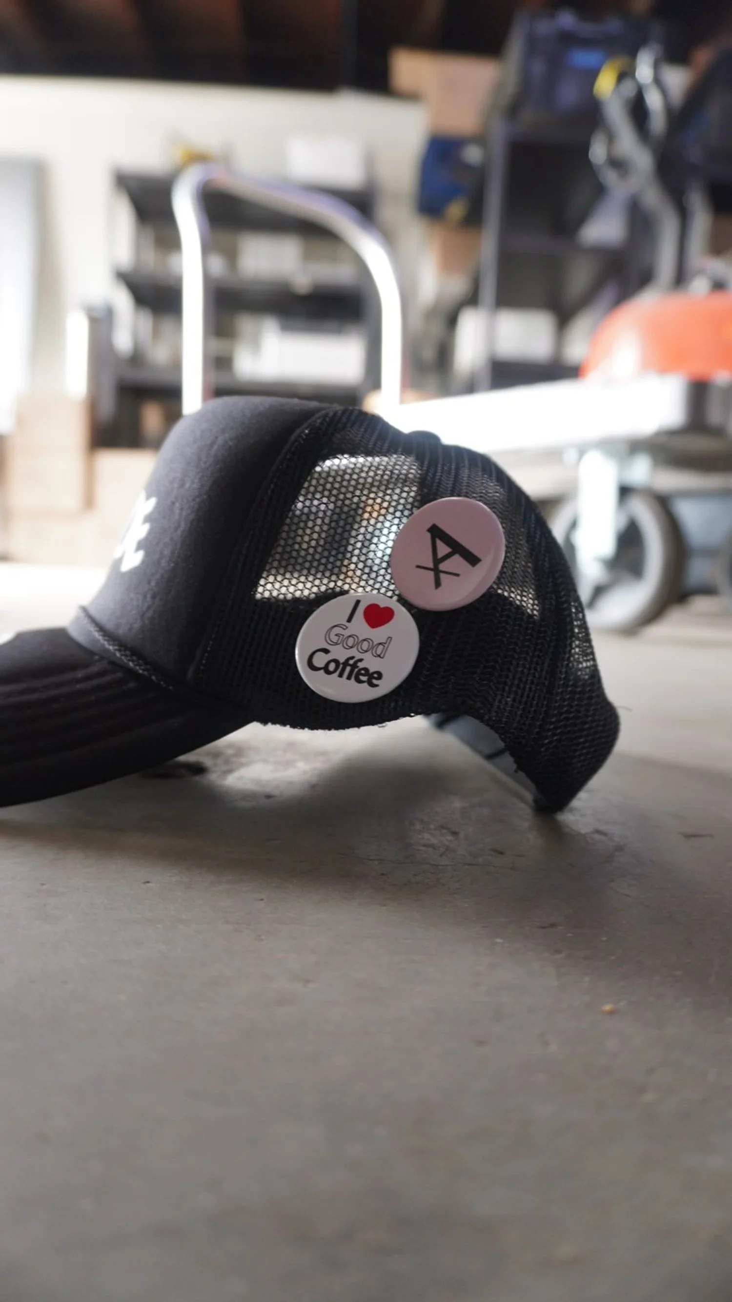 Close-up of a black baseball cap with two buttons on the side, one with a '4' symbol and another with 'I love good coffee' text and heart, resting on a concrete floor in a garage or workshop.