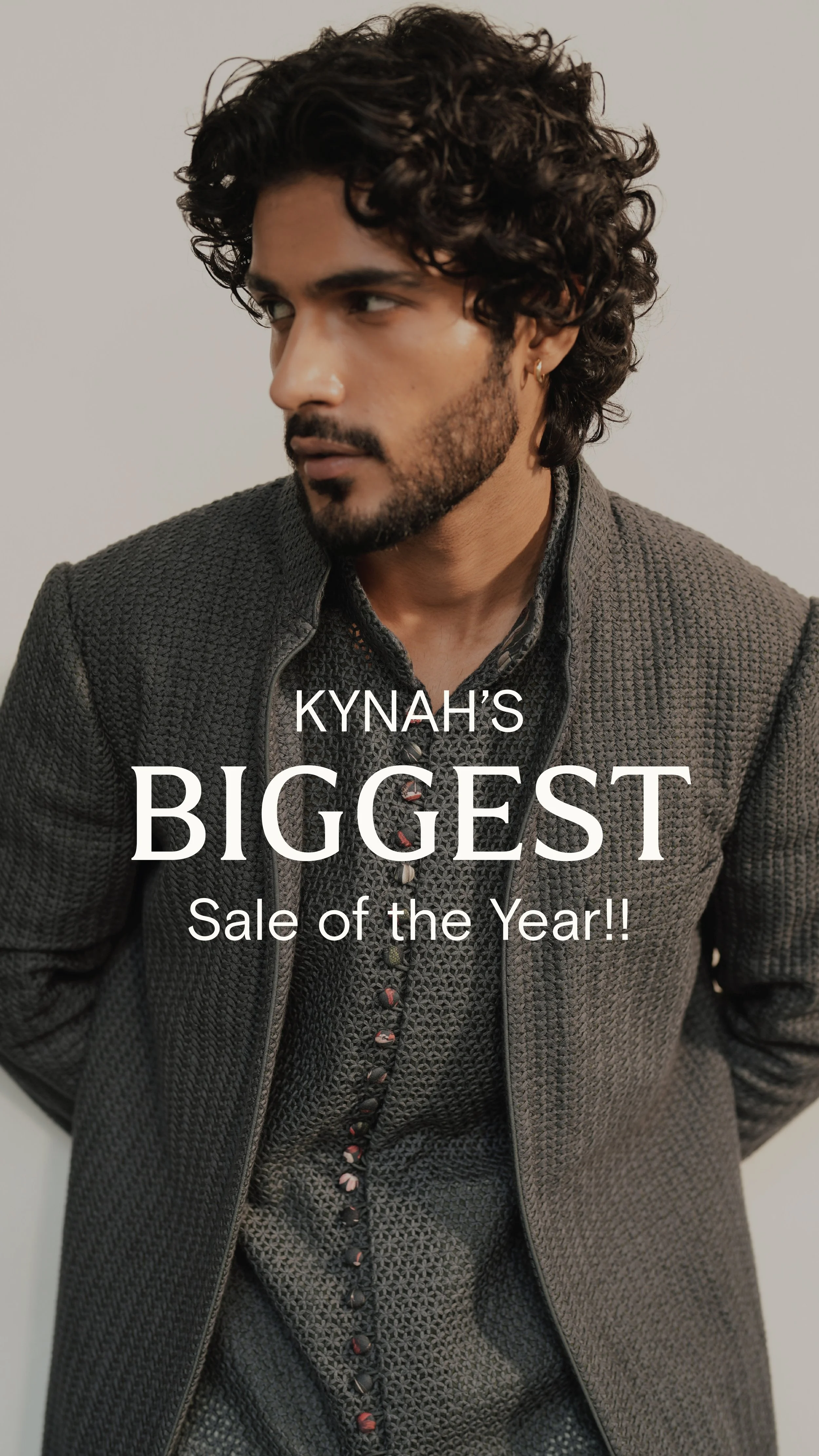 A man with dark curly hair and a beard, wearing a textured gray jacket and patterned shirt, looking to the side, with text overlay promoting Kynah's biggest sale of the year.