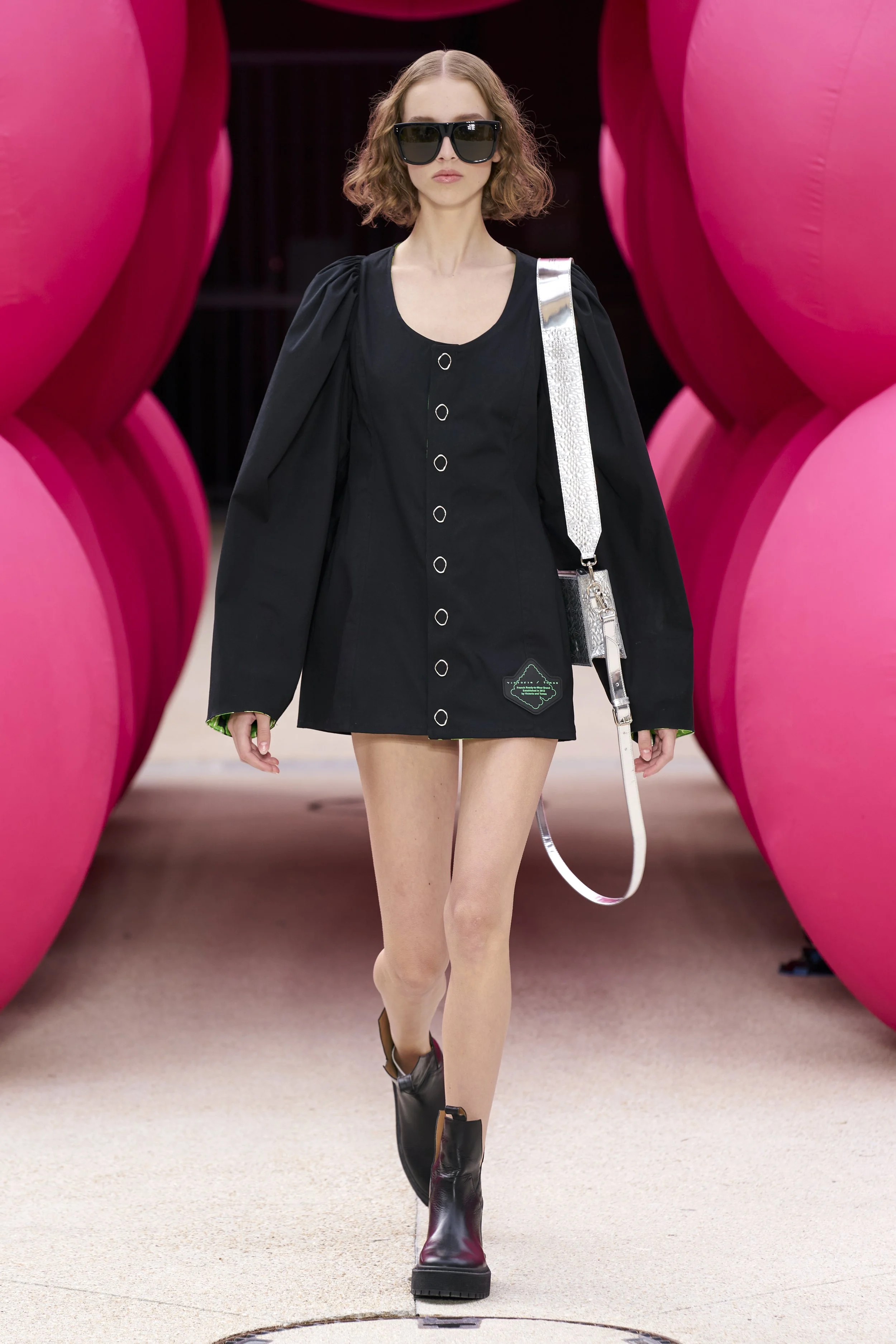 Model walking on runway wearing a short black dress with large sleeves, black sunglasses, and black combat boots, against a pink balloon installation backdrop.