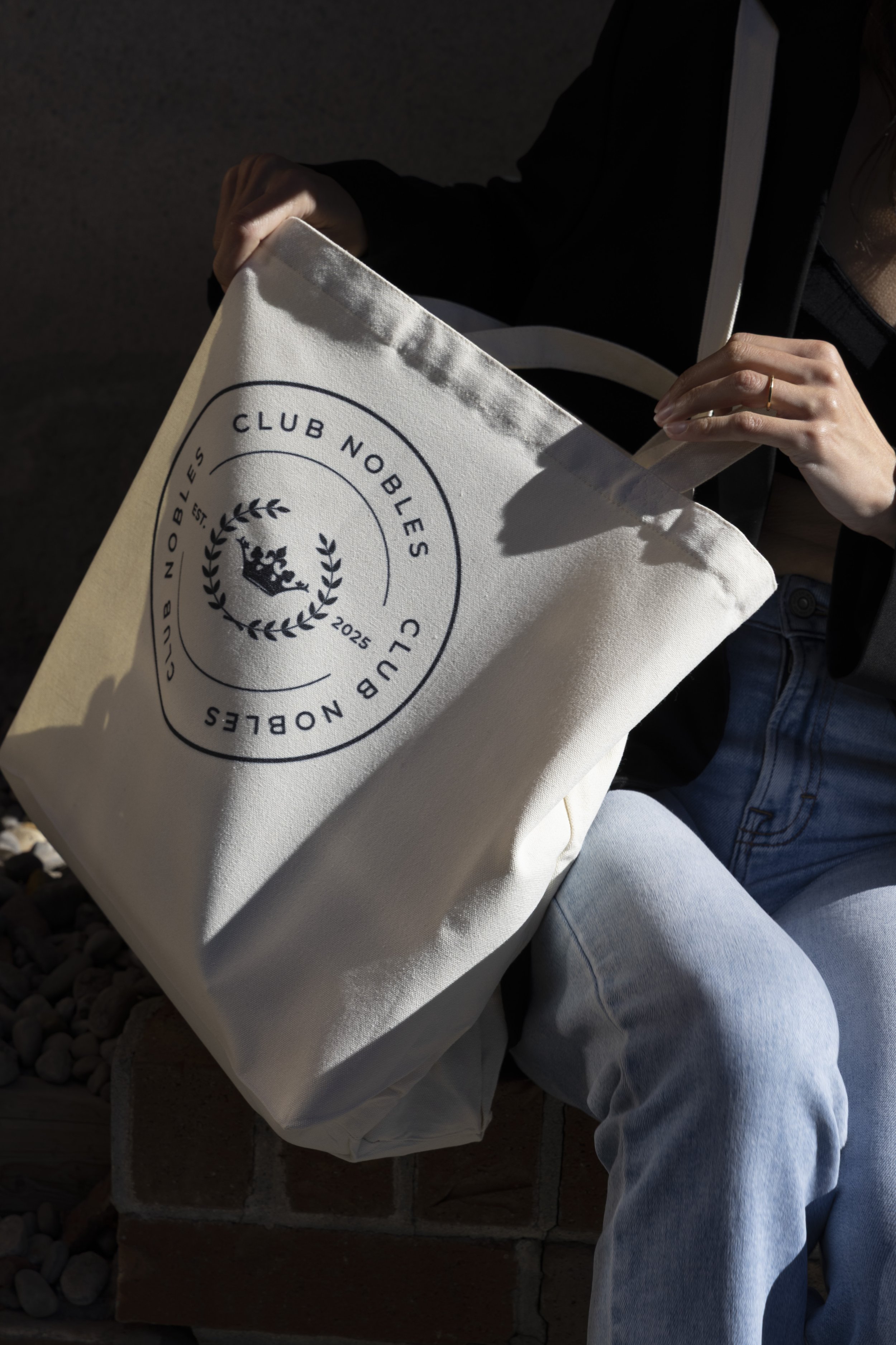 A person sitting outdoors holding a beige tote bag with a logo that reads "Club Nobles" with a crown and laurel wreath, and a date "2025".