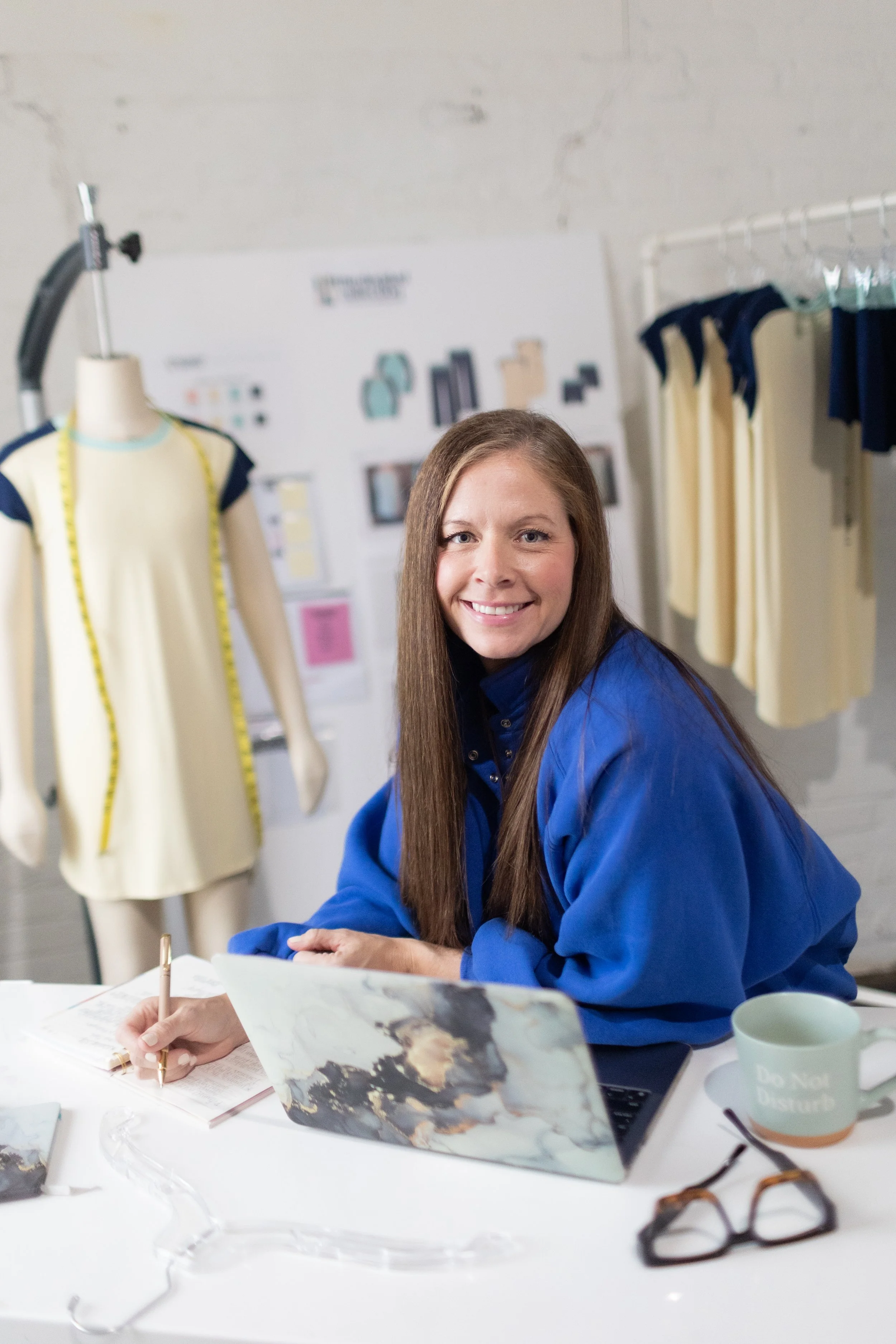 This is an image of Amanda Rango of ARD Fashion Consulting sitting in front of her computer. She is smiling at the camera, whilst writing on a notebook. In the background hangs a client's line of apparel and moodboard.