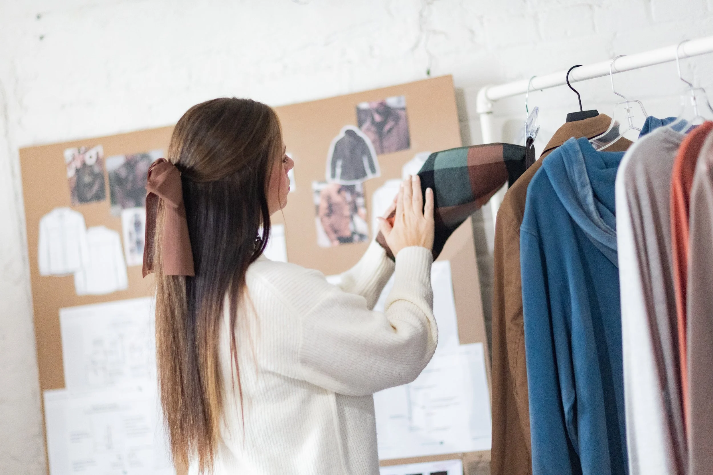 This is an image of Amanda Rango of ARD Fashion consulting interacting with a fabric swatch on a clothing rack with a variety of hanging samples. In the background is a cork board with blurred pinned inspiration photos