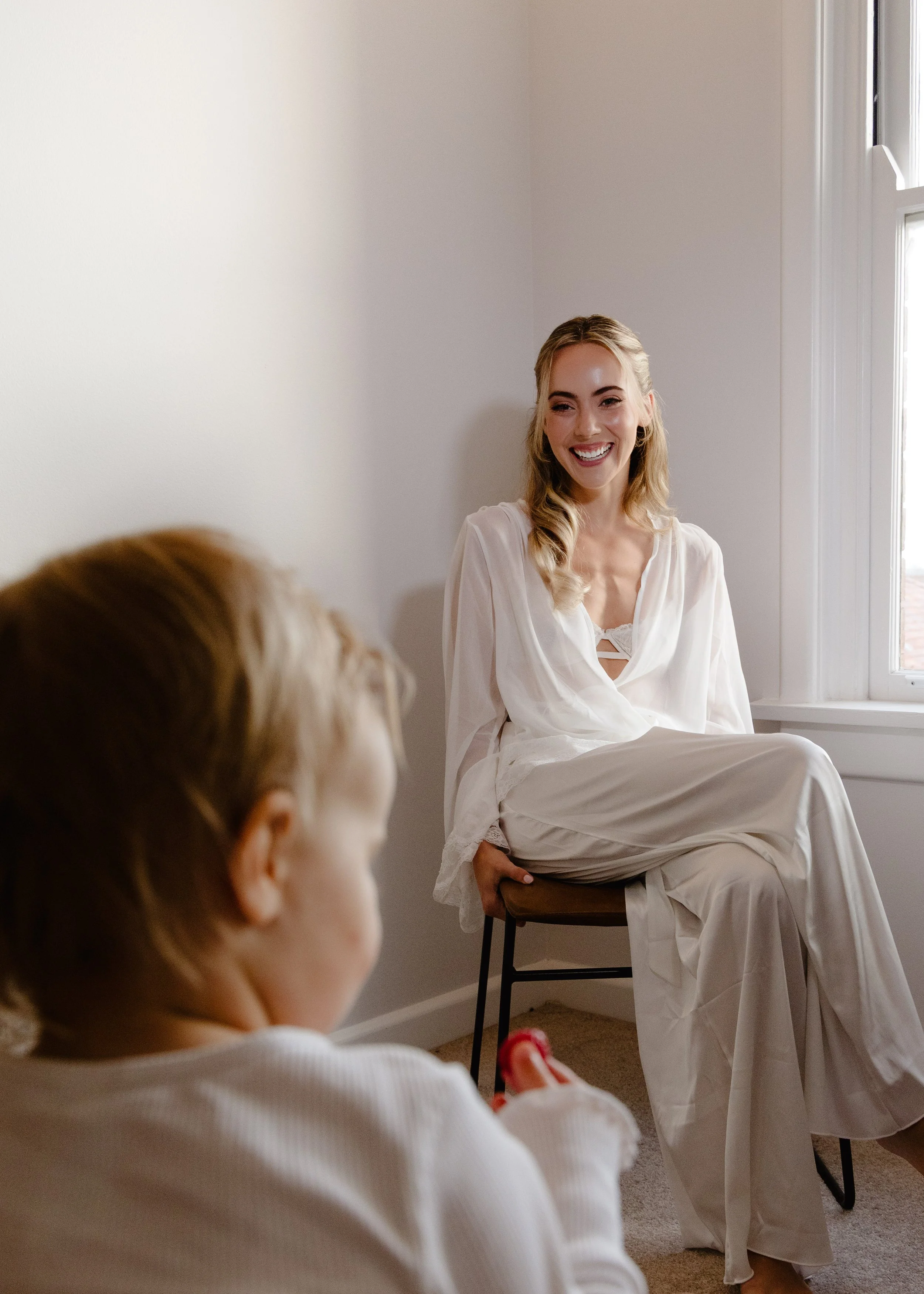 A woman in a white dress sitting on a chair by a window, smiling, while two children in white clothes are in the foreground, one holding a toy fork.