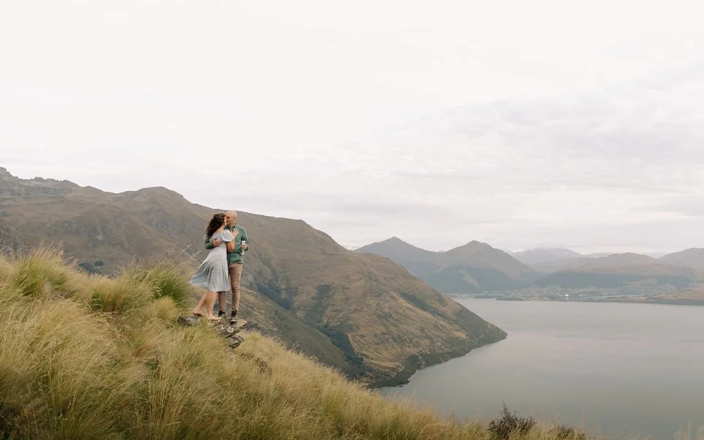 A couple standing on a grassy hill by a lake with mountains in the background, embracing and holding drinks.