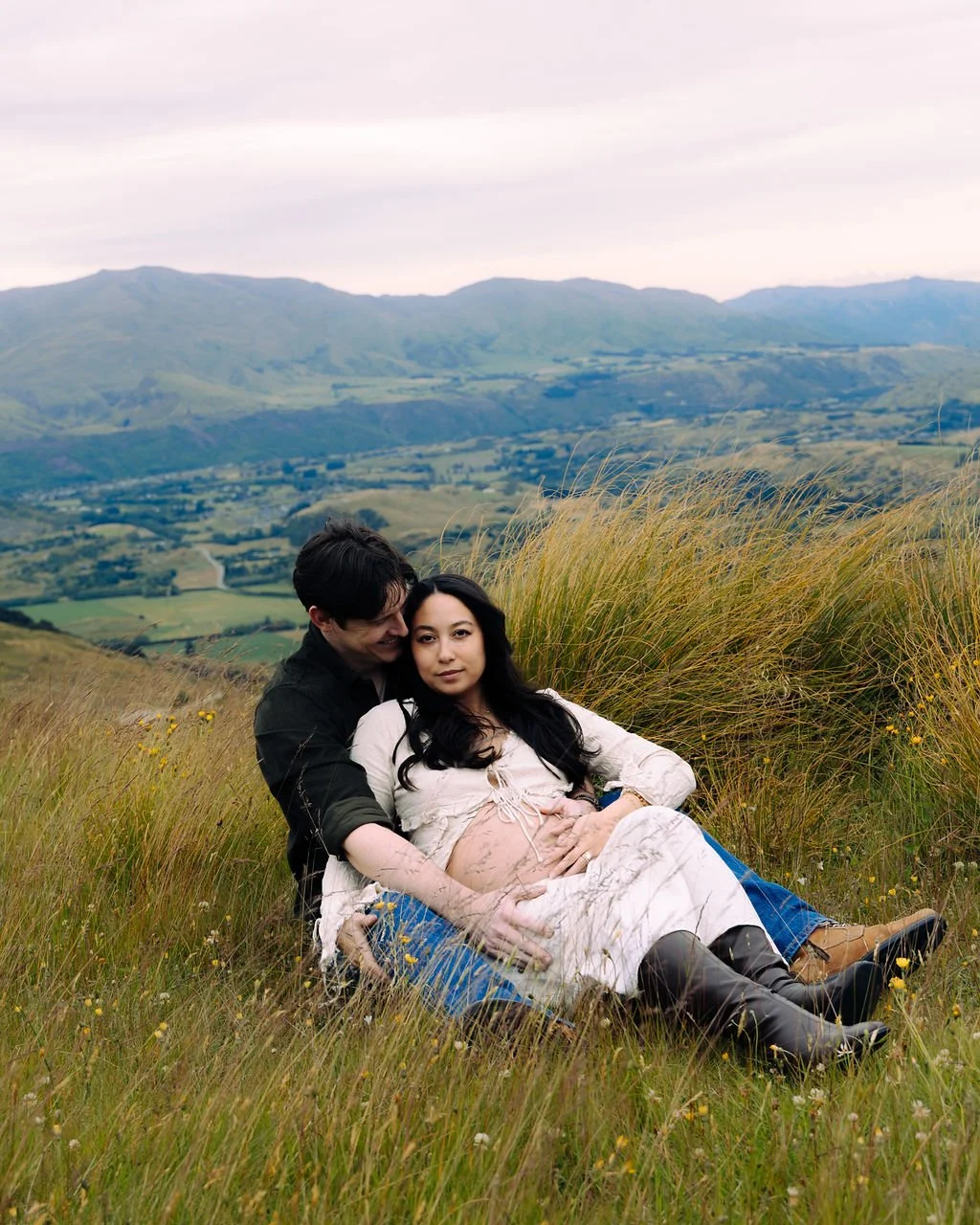 A pregnant woman and a man sit in a grassy field with rolling hills and mountains in the background.