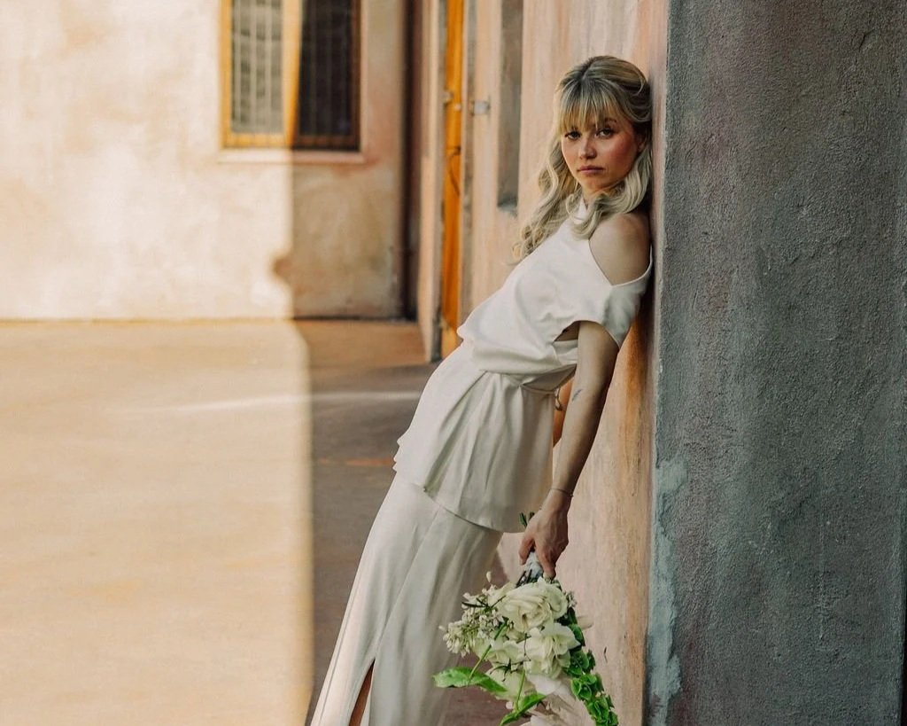 A woman in a white dress holding a bouquet of white flowers, leaning against a wall in an urban setting.