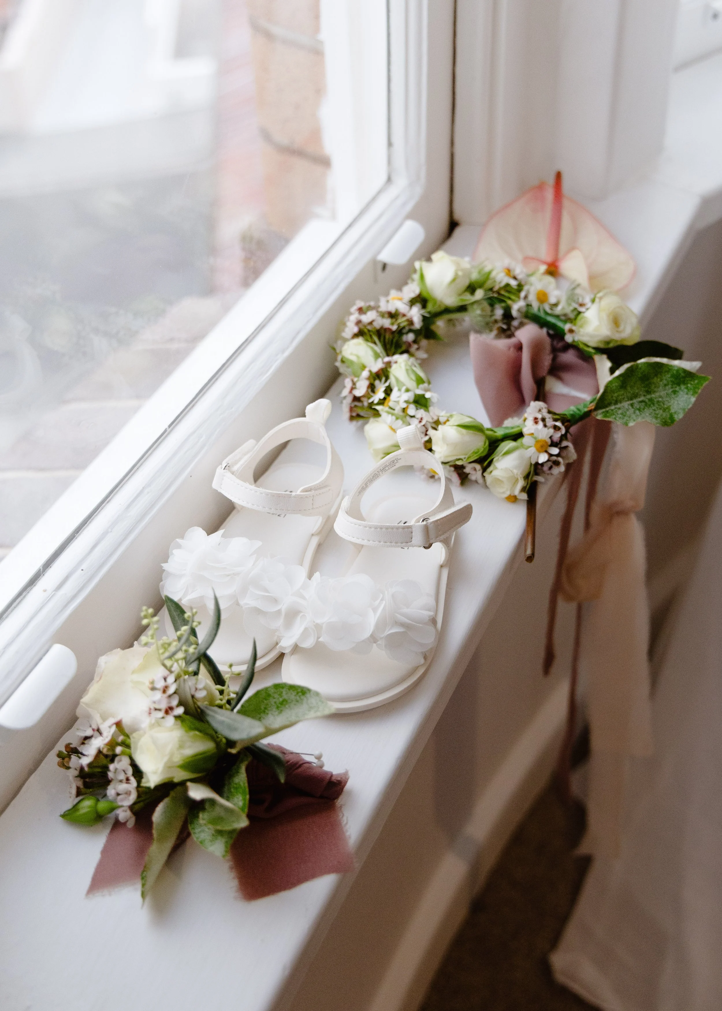Close-up of a flower crown, white sandals with white floral accents, and other floral decorations on a windowsill.