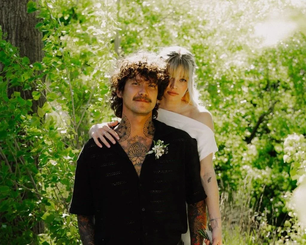 A man and woman standing outdoors in a sunny, green, wooded area, with the woman resting her arms on the man's shoulders.
