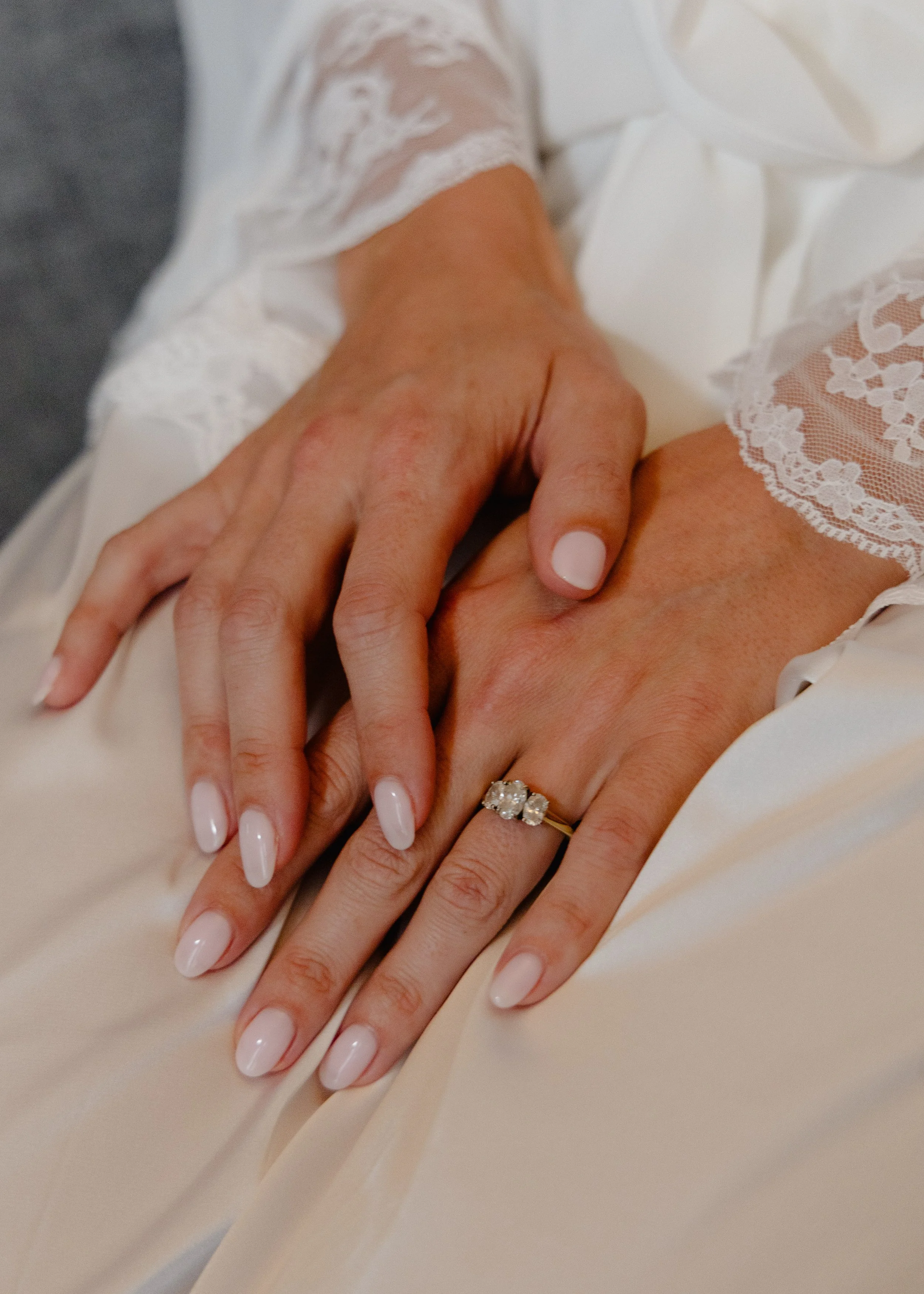 Close-up of a person’s hands with manicured nails resting on a light-colored fabric. The person is wearing a ring with a large gemstone, and is dressed in white with lace details.