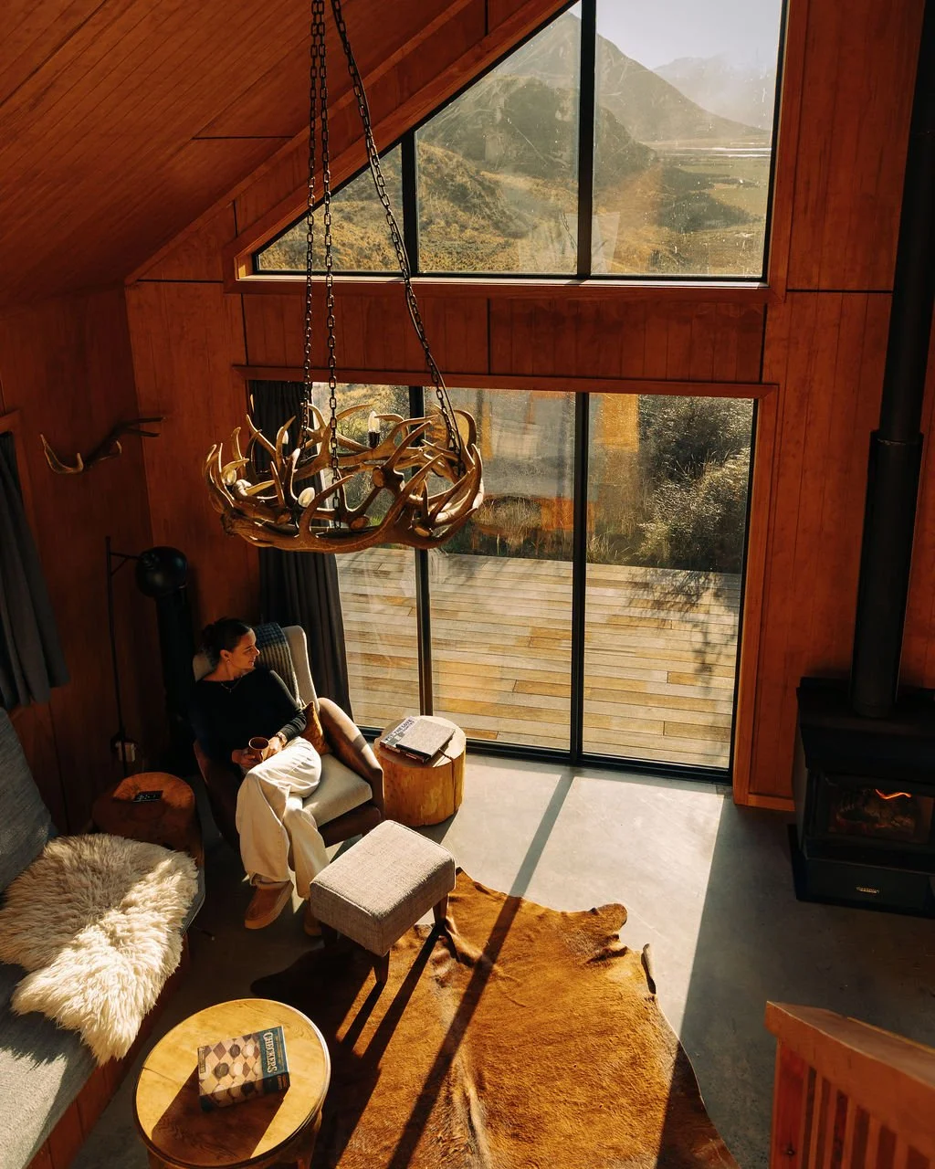 A cozy living room with wooden walls and large windows showing a mountain view. A woman is sitting in a chair holding a cup, with a sheepskin blanket on a nearby sofa, a cowhide rug on the floor, and a round coffee table.