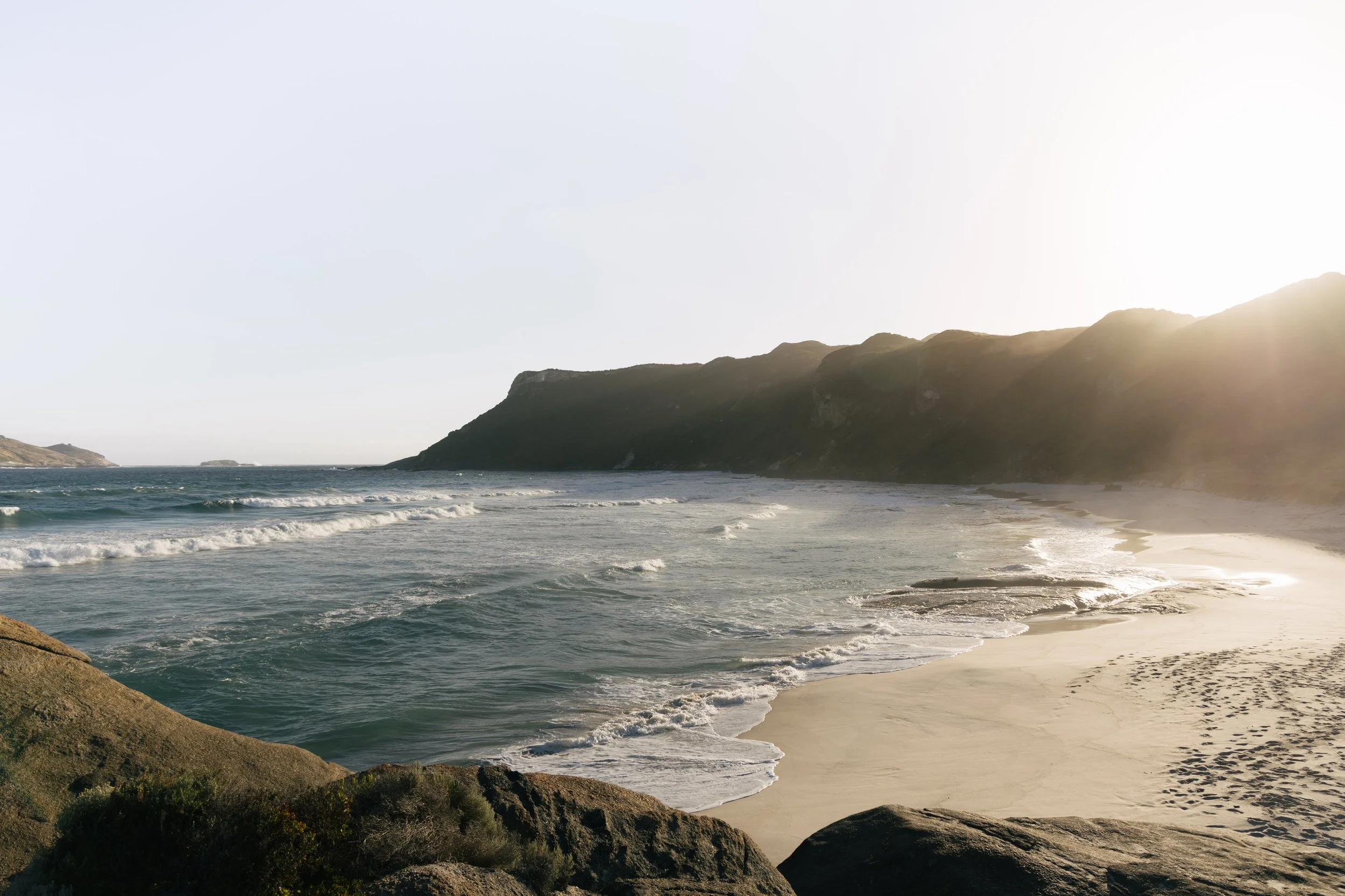 Sunset over the ocean with waves crashing onto the sandy beach, cliffs in the distance, and rocks in the foreground.