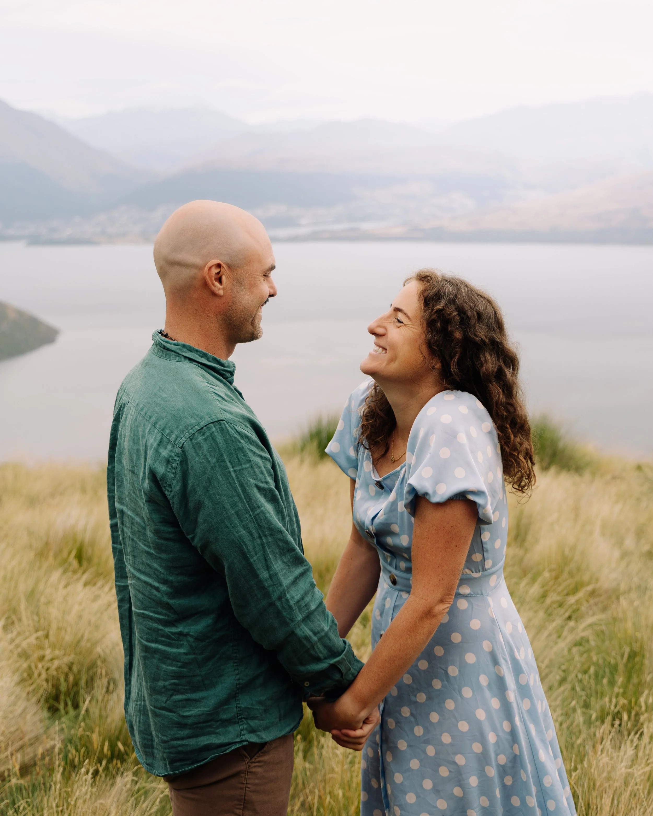 A smiling couple holding hands and facing each other outdoors in a field with mountains in the background.