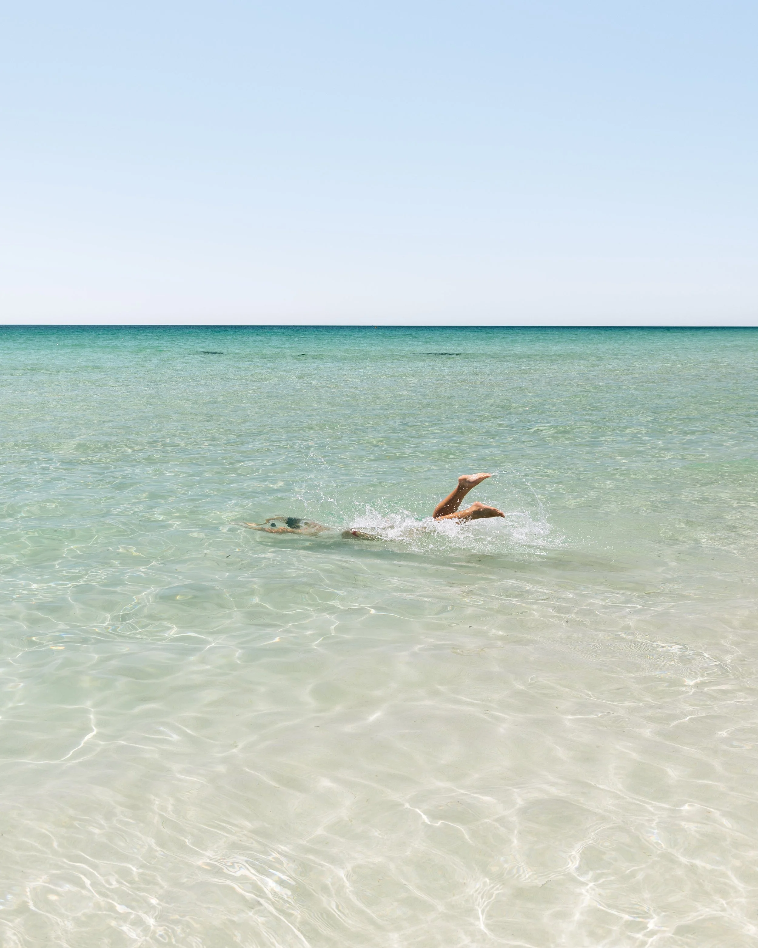 A person swimming in clear, shallow ocean water near the shore. The swimmer's legs are visible above the water, and the ocean continues into the horizon under a clear blue sky.