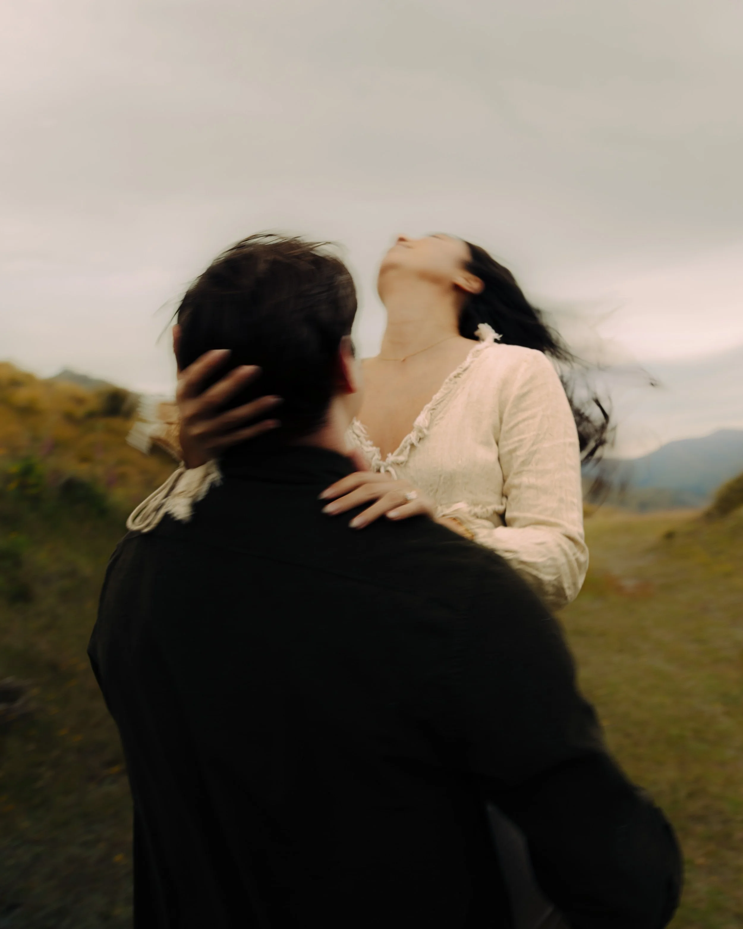 A woman and a man are embracing each other outdoors on a cloudy day. The woman is tilting her head back and has her eyes closed, while the man's face is hidden as he holds her. The background shows a blurry landscape with mountains and greenery.
