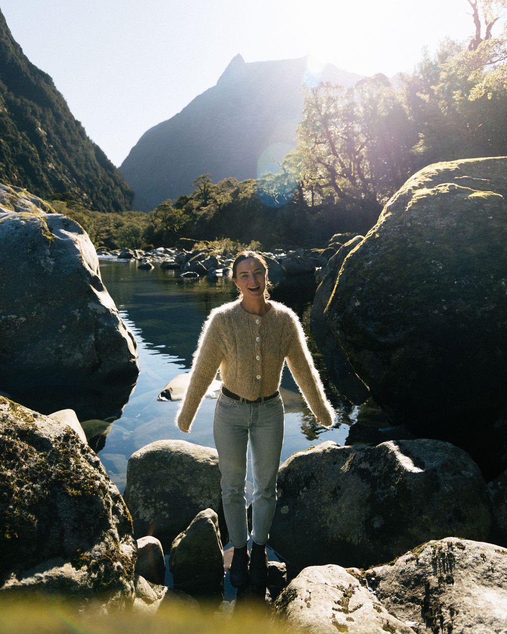 A woman standing on rocks in a river valley surrounded by large boulders, trees, and mountains with the sun shining brightly overhead.