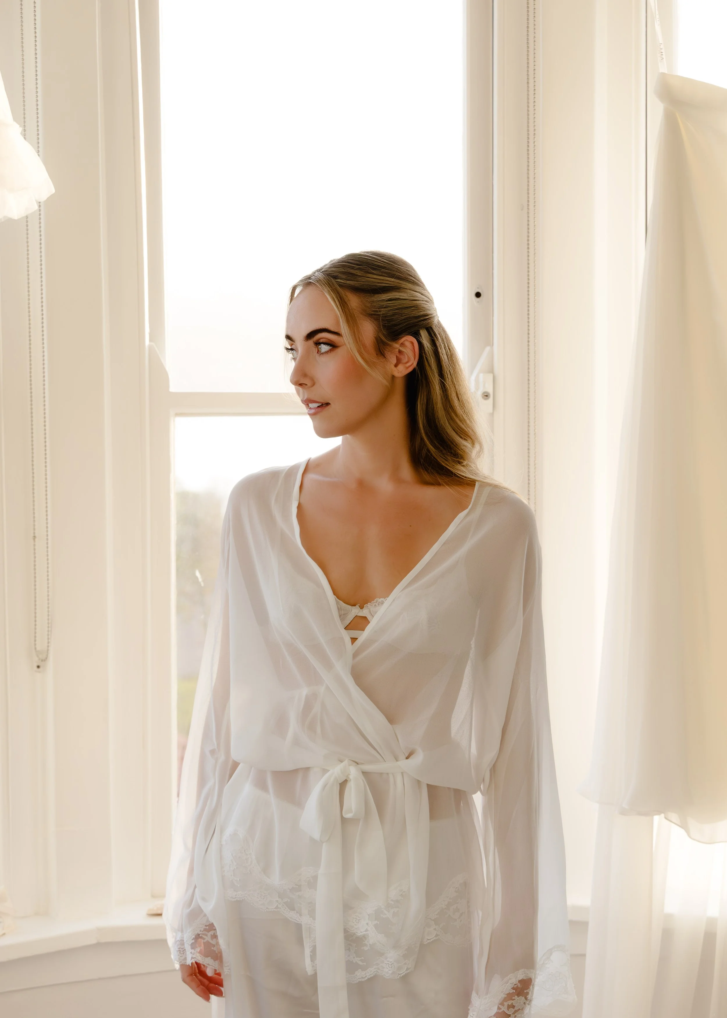 A woman with blonde hair styled in loose waves, wearing a white satin robe with lace trim, standing in front of a bright window with white curtains.