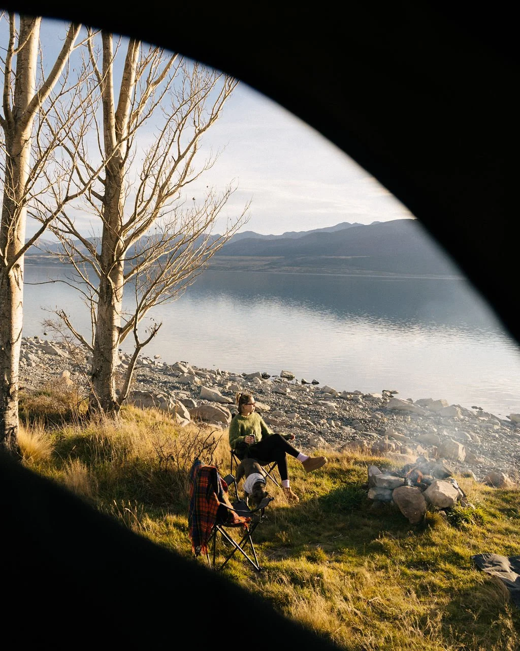 Person relaxing by a lakeside campfire with a chair, trees, rocks, and mountains in the background, viewed through a car window