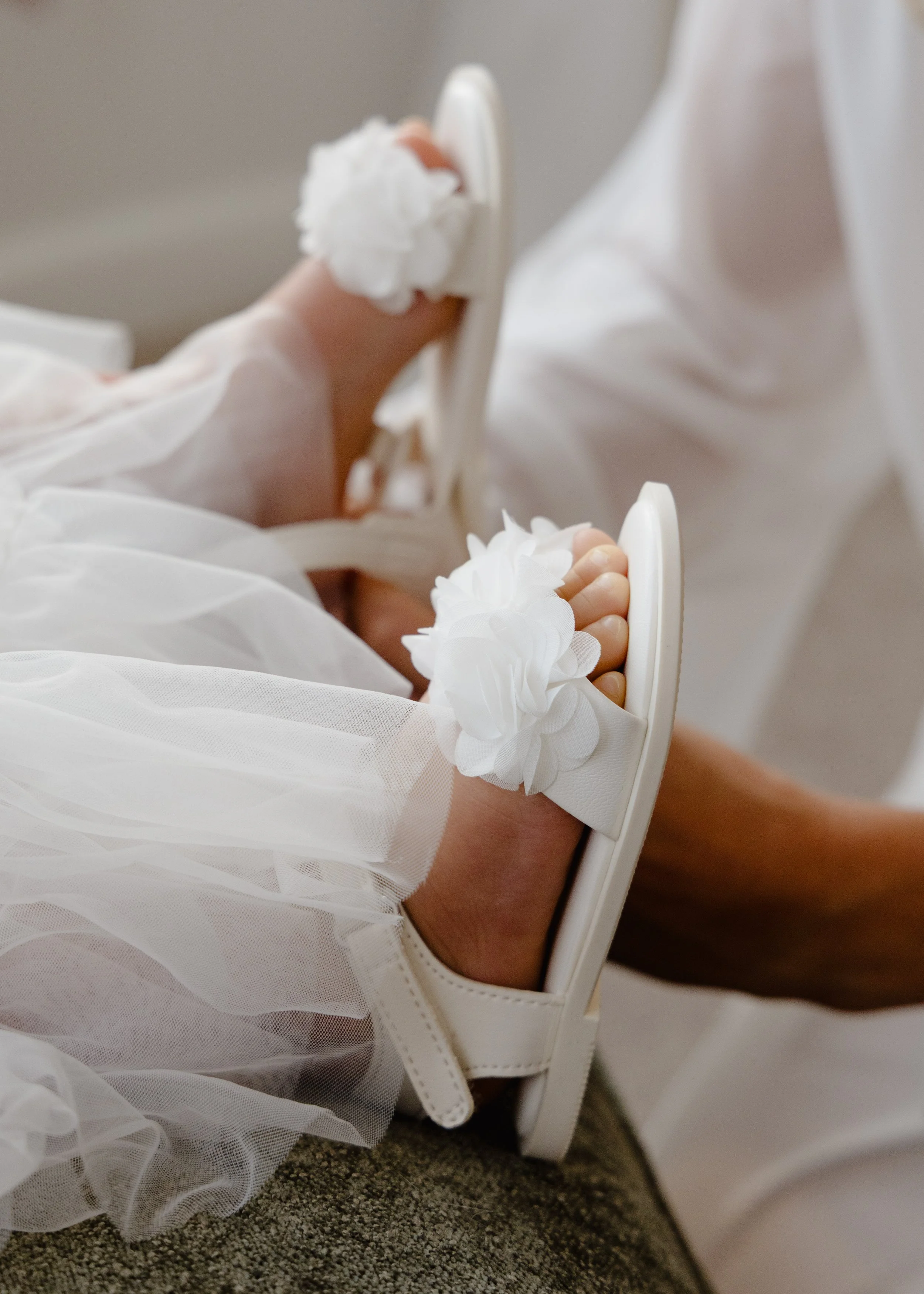 Close-up of a newborn baby's hand gripping a white hospital bed rail, dressed in a white garment with ruffled sleeves, on a gray carpet.