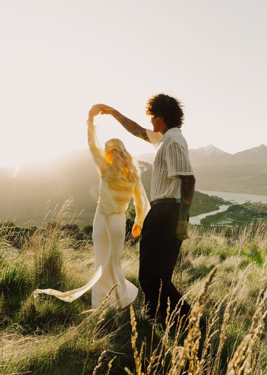 A couple dancing in a grassy field during sunset, with mountains, a river, and a lake in the background.