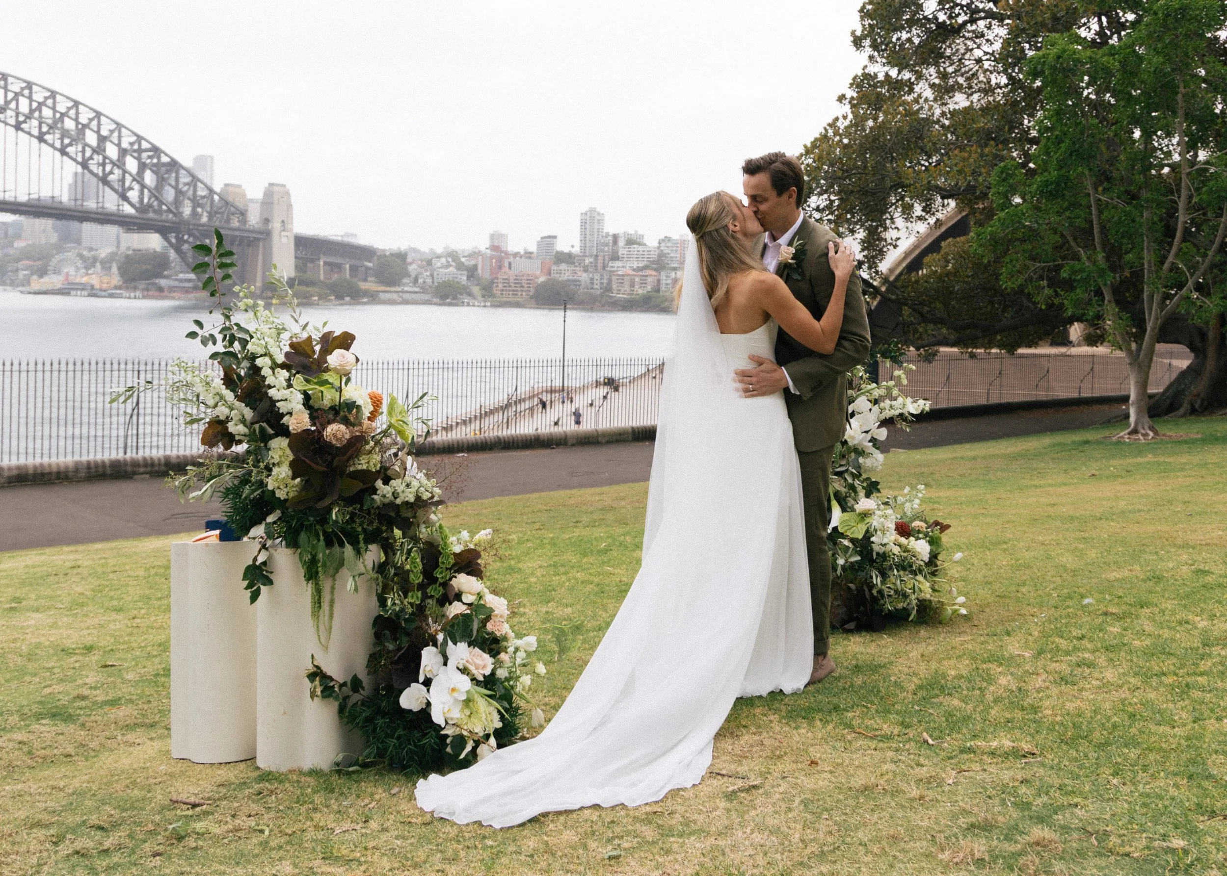 A couple sharing a kiss at their wedding ceremony near a body of water, with a city skyline and bridge in the background. There are floral arrangements and a white cloth on the ground.