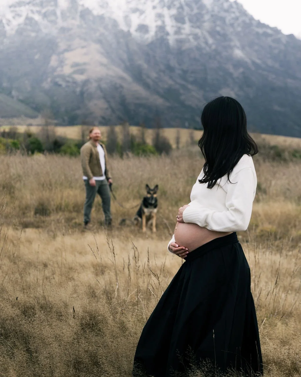 A pregnant woman with black hair in a white sweater and black skirt standing in a field, looking at a man and a dog in the background with mountains behind them.