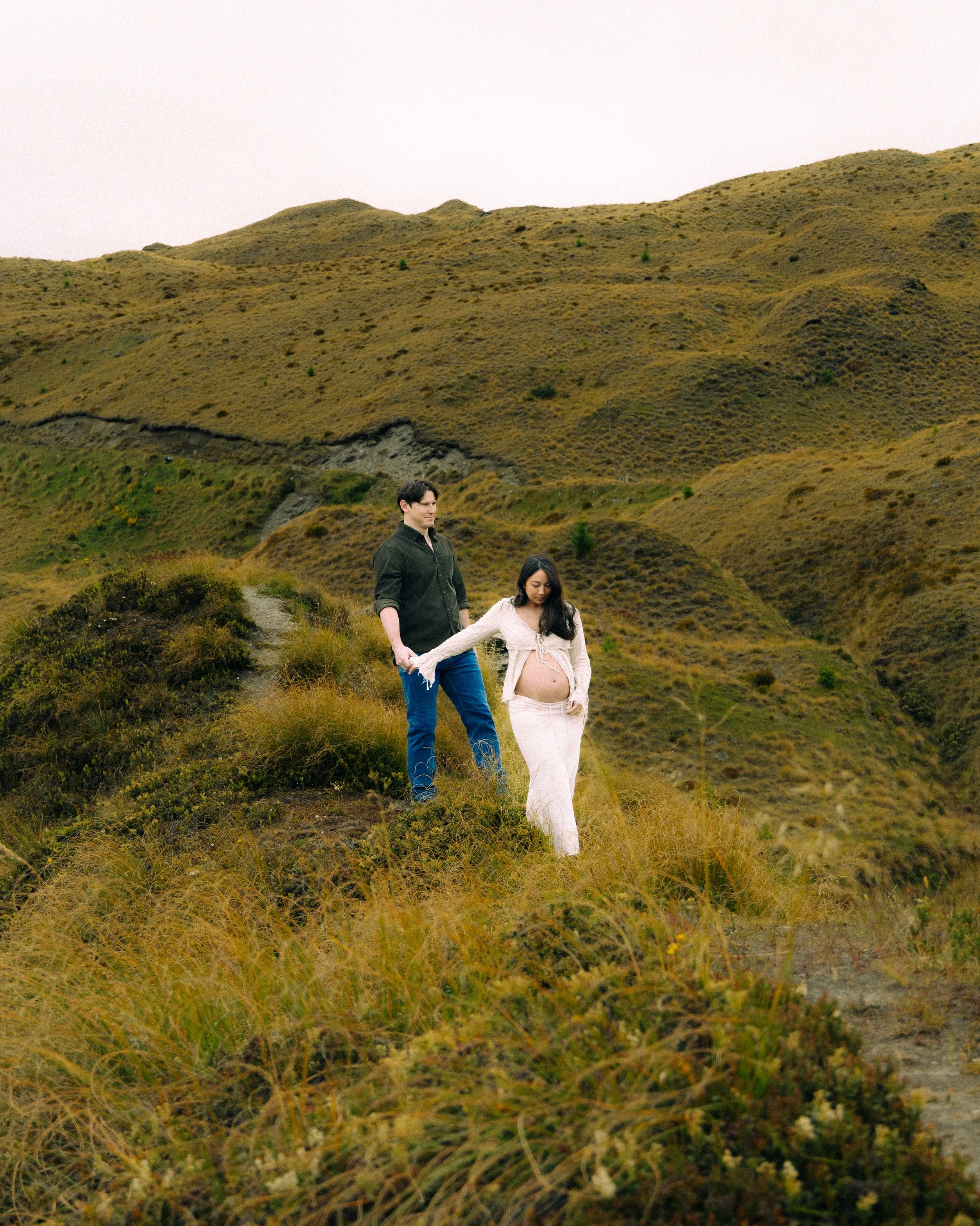 A man and a pregnant woman holding hands walking down a trail in a grassy hillside landscape.