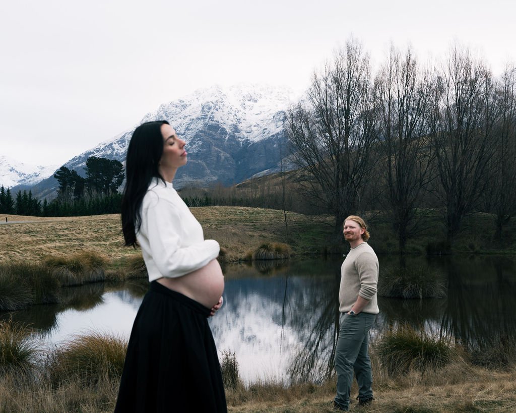 A pregnant woman with dark hair and light skin standing by a pond in a natural landscape, with a man with light skin and reddish hair looking at her, snow-capped mountains and leafless trees in the background.