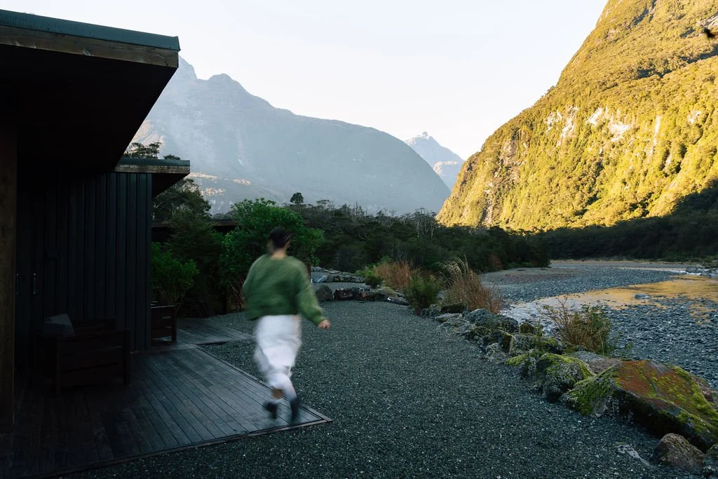 Person walking near a river valley with mountains in the background, next to a dark-colored building in a scenic outdoor setting.