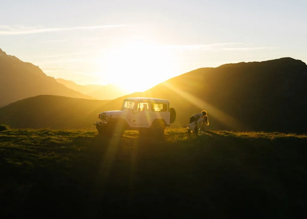 A silhouette of a vintage off-road vehicle and a person near a couple of dogs on a grassy hill during sunset, with mountains in the background.