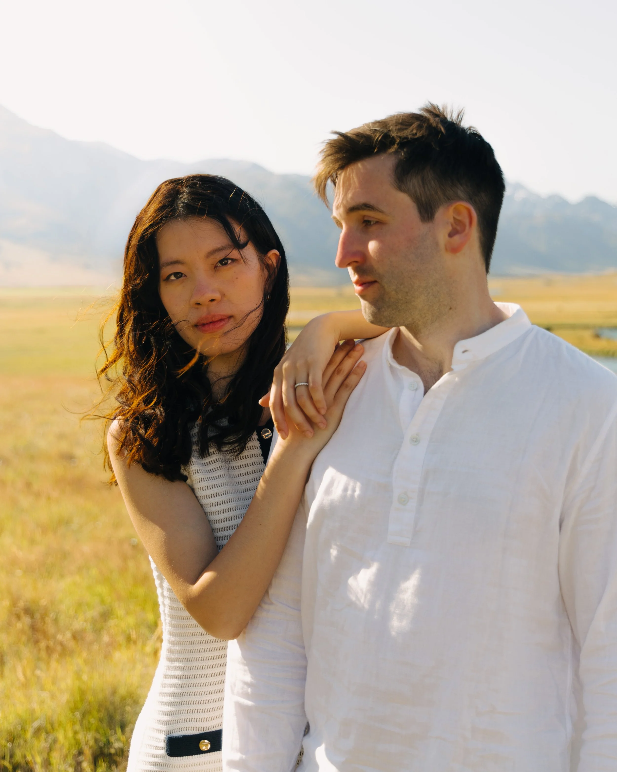 A closer view of a young woman and a young man standing outdoors in a field, with mountains in the background, during daylight. The woman has long, dark hair and is looking at the camera. The man has short, dark hair and is looking at the woman. The 