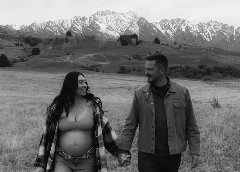 A couple enjoying a walk in an open field with mountains in the background, smiling and holding hands.