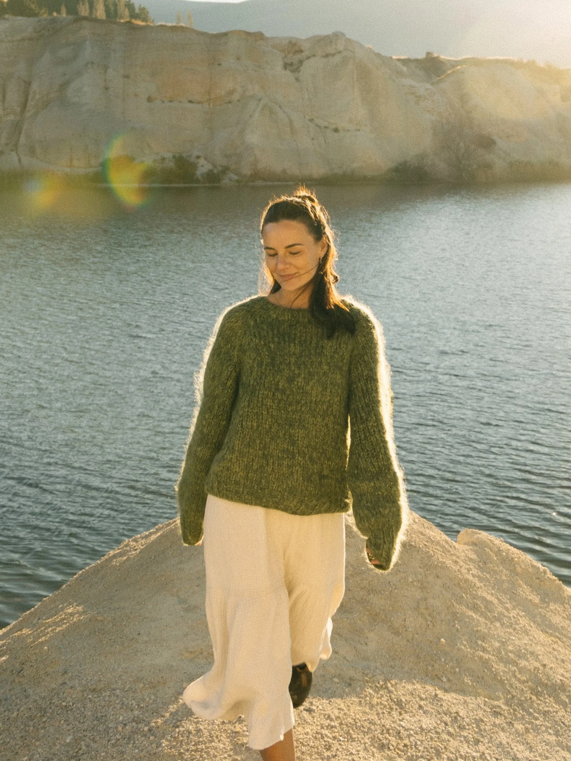 A woman in a green sweater and white skirt walking along rocky coast at sunset, with cliffs and water in the background.