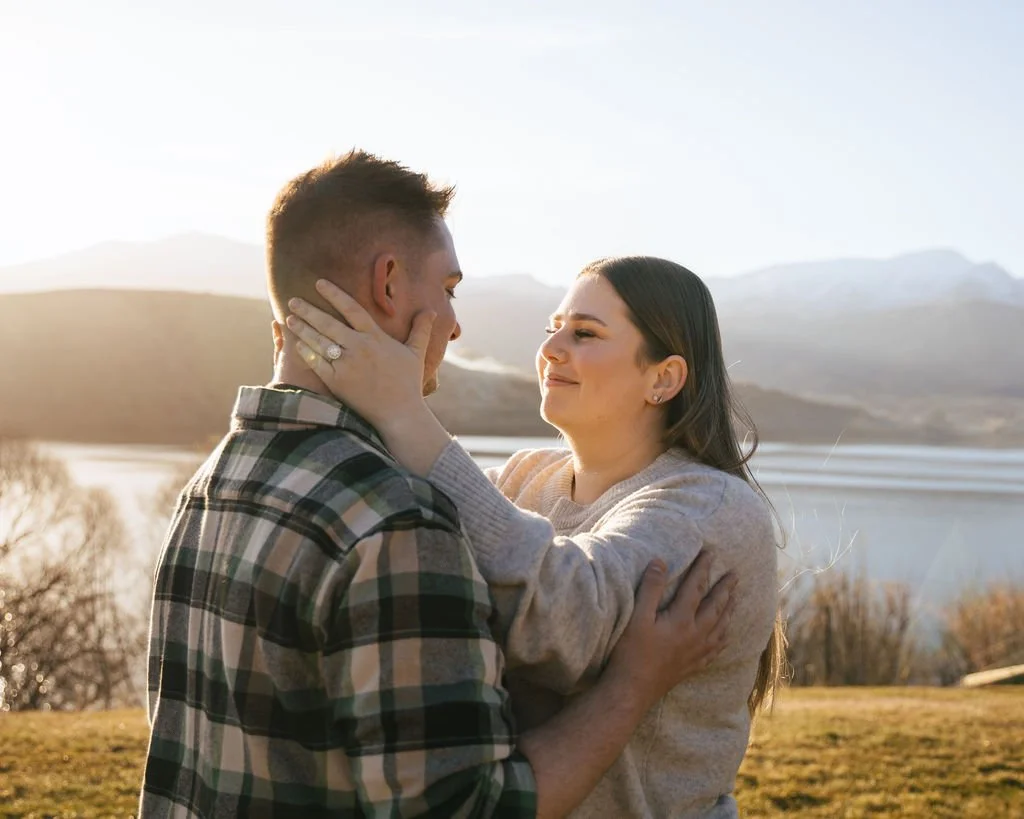 A couple embracing outdoors during sunset with mountains and a lake in the background.